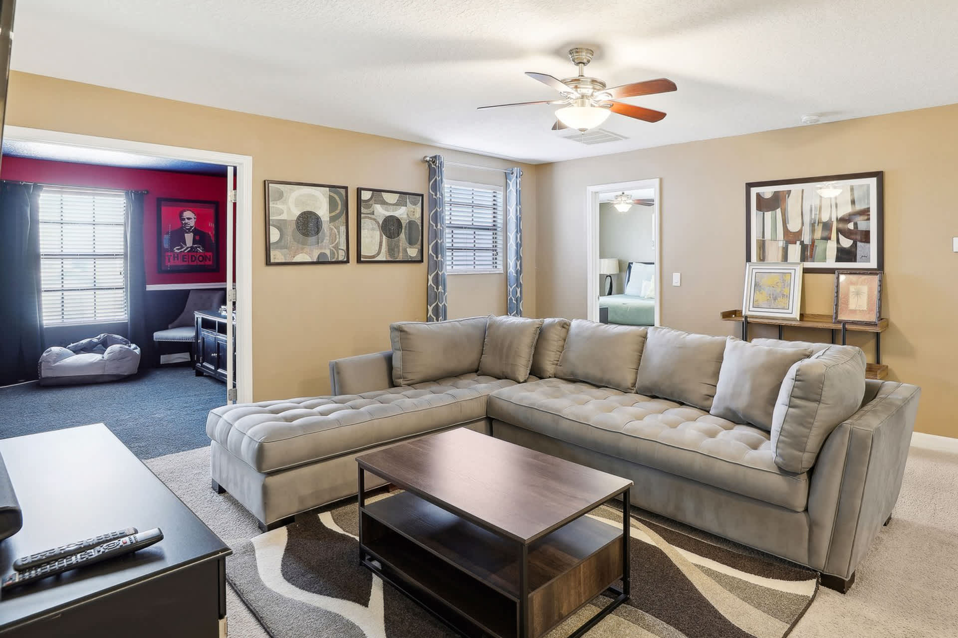 Cozy upstairs media room with a large gray sectional sofa and a dark wood coffee table on a patterned area rug. A doorway leads to another room, and framed artwork adorns the walls.