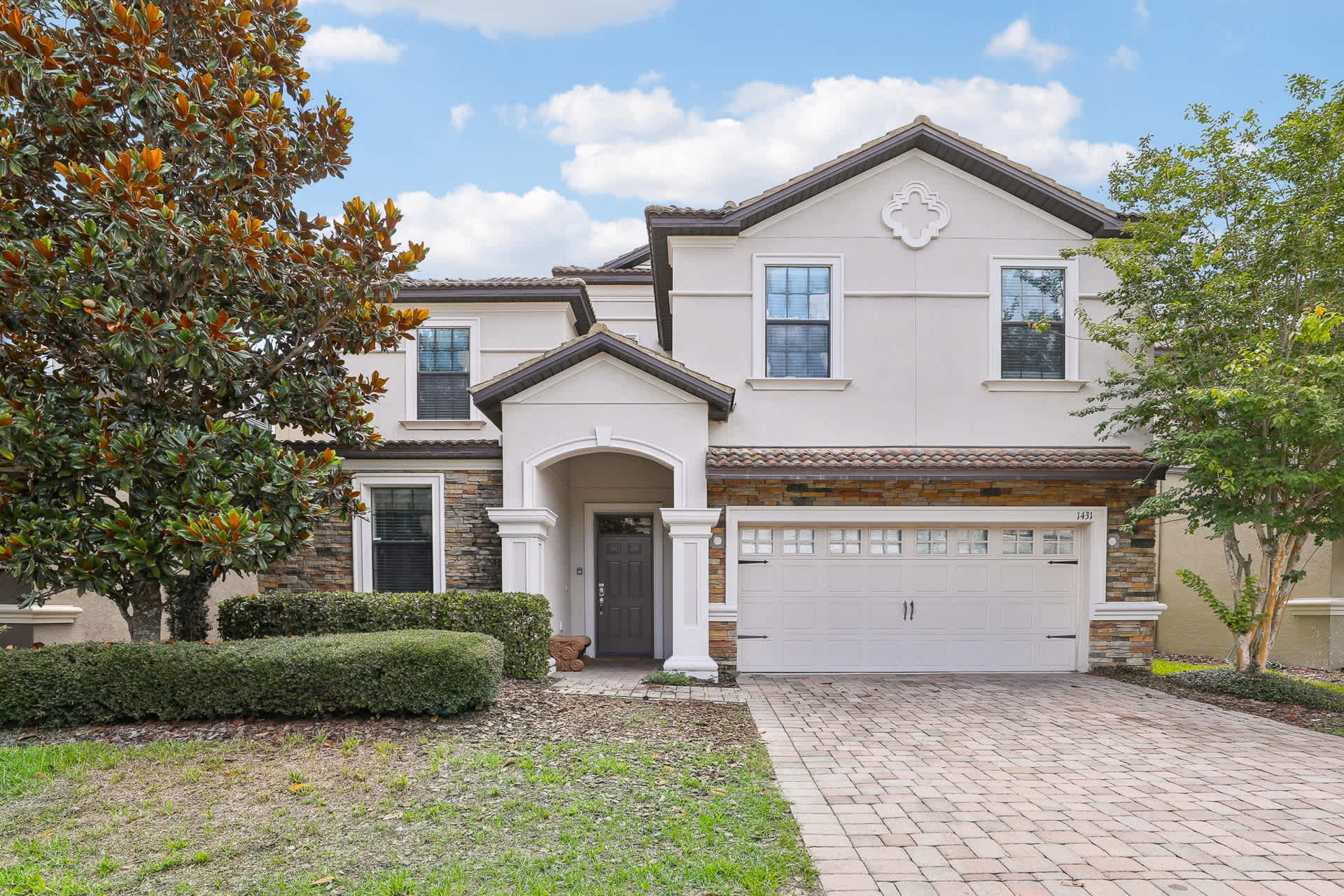 Two-story house with a light-colored stucco and stone facade, featuring a two-car garage and a covered entryway. A paved driveway leads to the garage, and mature trees and shrubs adorn the front yard.