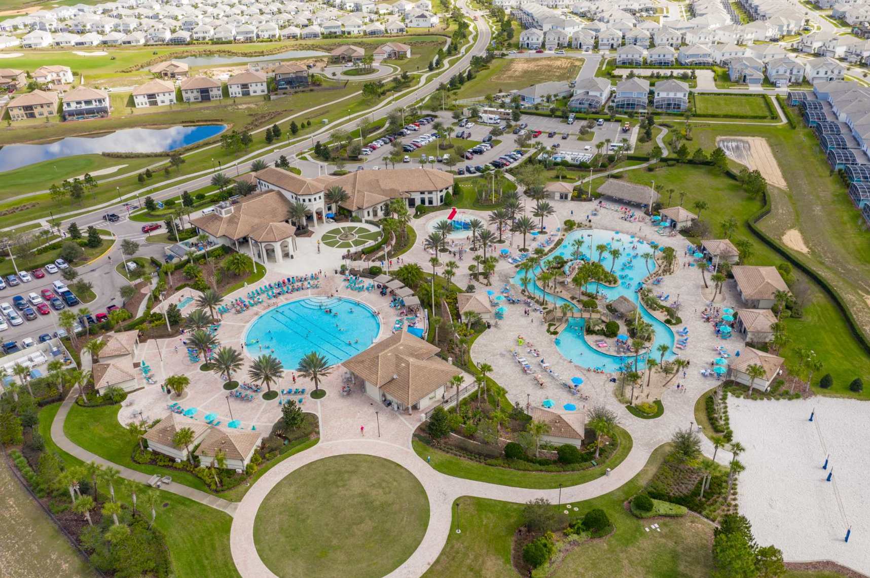 An aerial view captures a large resort-style communal pool complex with multiple pools, palm trees, cabanas, and lounge areas, surrounded by residential homes and a golf course.