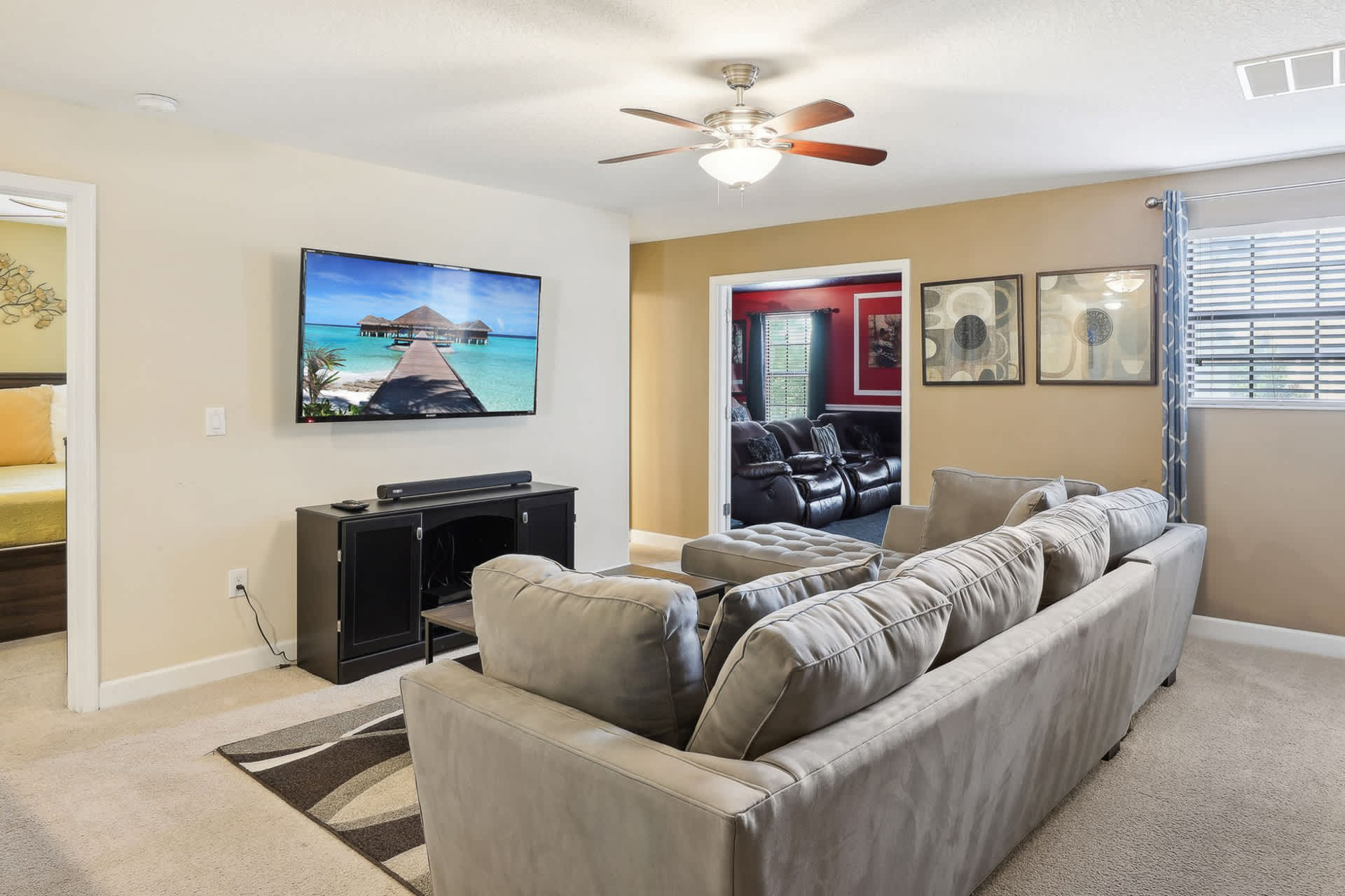Media room featuring a wall-mounted flat-screen TV above a dark media console, with a large gray sectional sofa providing ample seating. A doorway leads to a red-walled room, likely a cinema or game room.