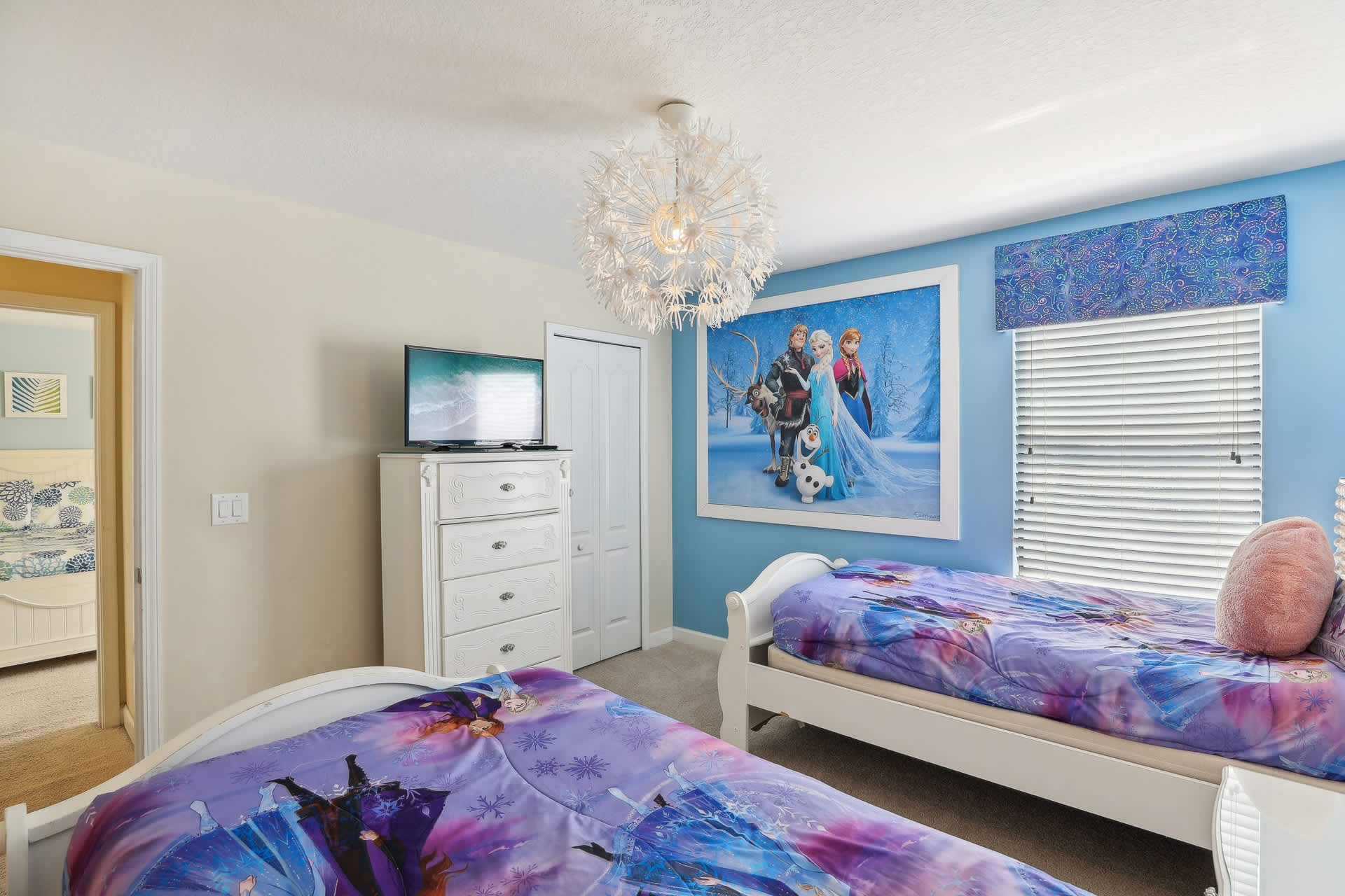 Kids' themed bedroom with two white twin beds featuring 'Frozen' bedding, a large 'Frozen' wall mural, a white dresser with a TV, and a light blue accent wall.