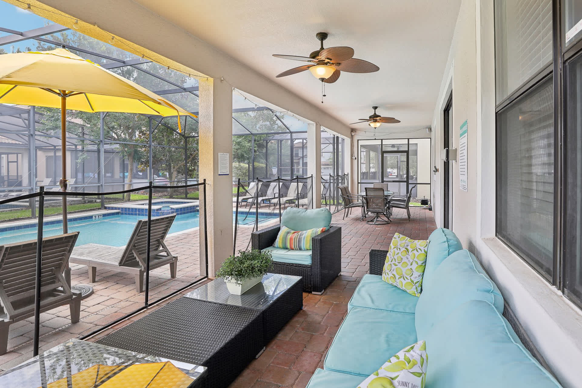 Covered outdoor patio area adjacent to the screened pool, furnished with a light blue sofa, an armchair, and a coffee table. Ceiling fans are installed overhead, and a dining table with chairs is visible further back.