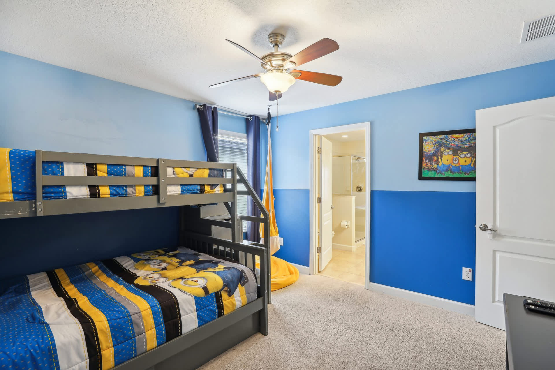 A colorful children's bedroom showcases a gray bunk bed with Minions bedding, blue two-tone walls, a ceiling fan, and an open door leading to an en-suite bathroom.