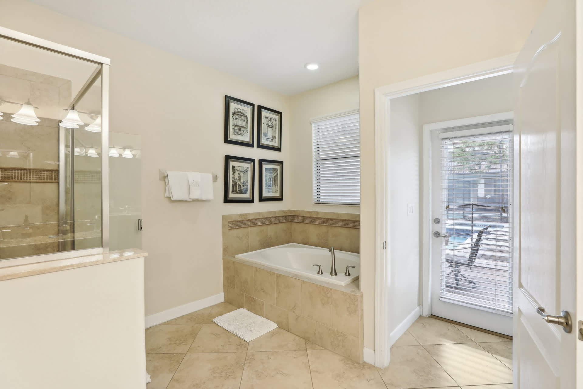 A corner of a light-colored bathroom featuring a built-in bathtub surrounded by tiled walls, a small white rug, and an open door leading to an outdoor patio with a pool.