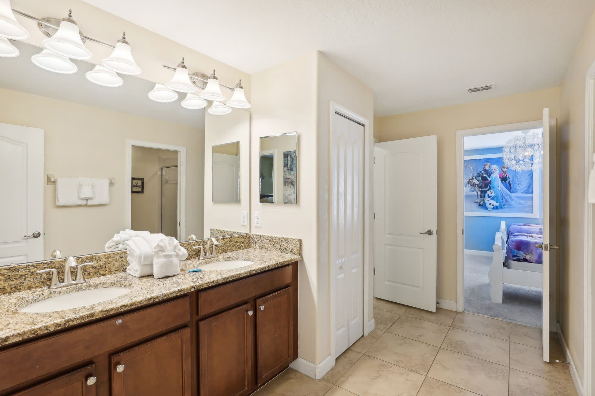 Bright bathroom featuring a double vanity with granite countertops, two large mirrors, white cabinets, and tiled flooring, with a glimpse of a themed bedroom through an open door.