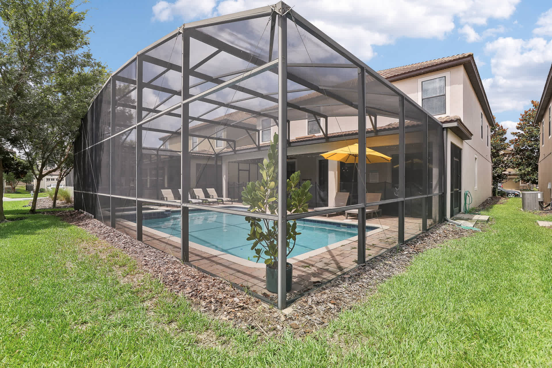 An outdoor private swimming pool is enclosed by a screen cage, surrounded by a paved deck with lounge chairs and a yellow umbrella, set against a backdrop of green grass and a two-story house.