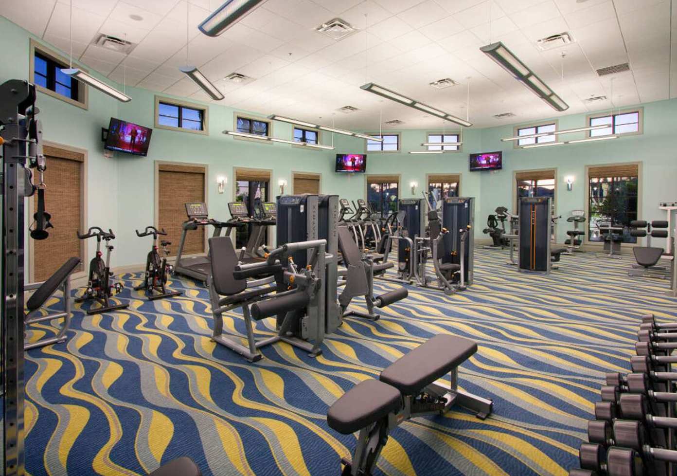 A spacious fitness center featuring a variety of modern exercise equipment, light blue walls, and a vibrant blue, yellow, and white wavy patterned carpet. Multiple flat-screen TVs are mounted on the walls.