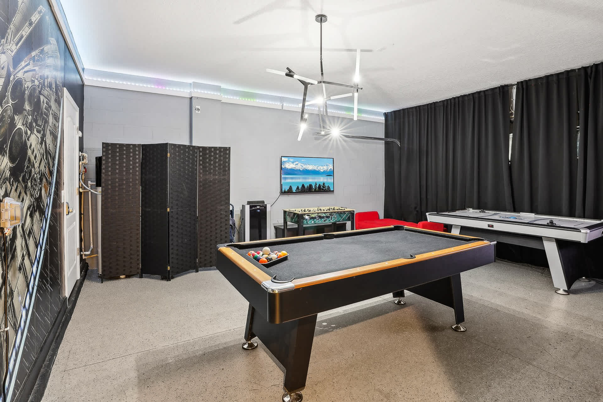 Spacious game room with gray walls and a large mural, featuring a black pool table, a foosball table, and an air hockey table. The room has modern lighting fixtures and dark curtains, providing a dedicated entertainment space.