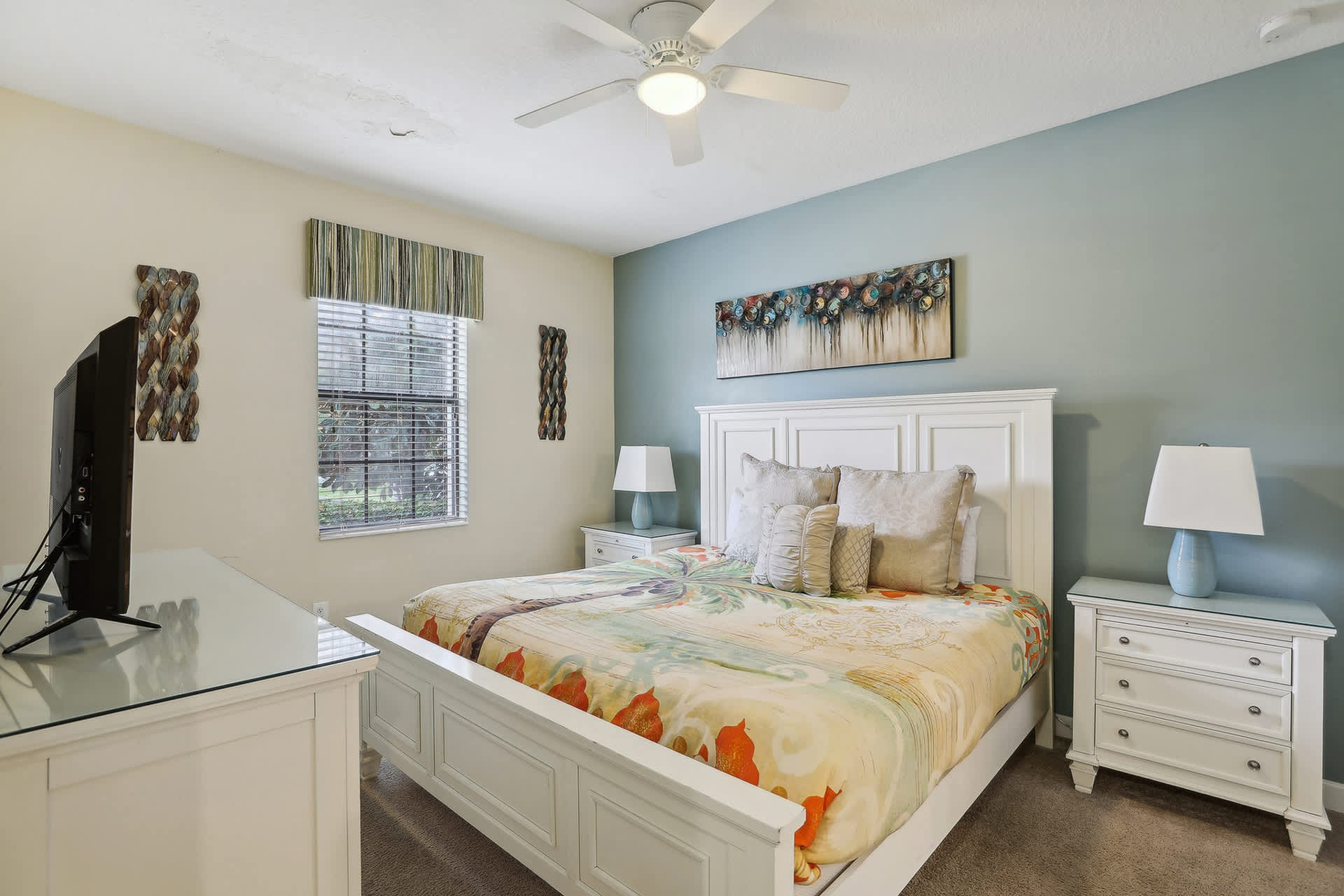 A bright bedroom with a white bed frame, colorful bedding, a light blue accent wall, two nightstands with lamps, and a ceiling fan, featuring a window with blinds.