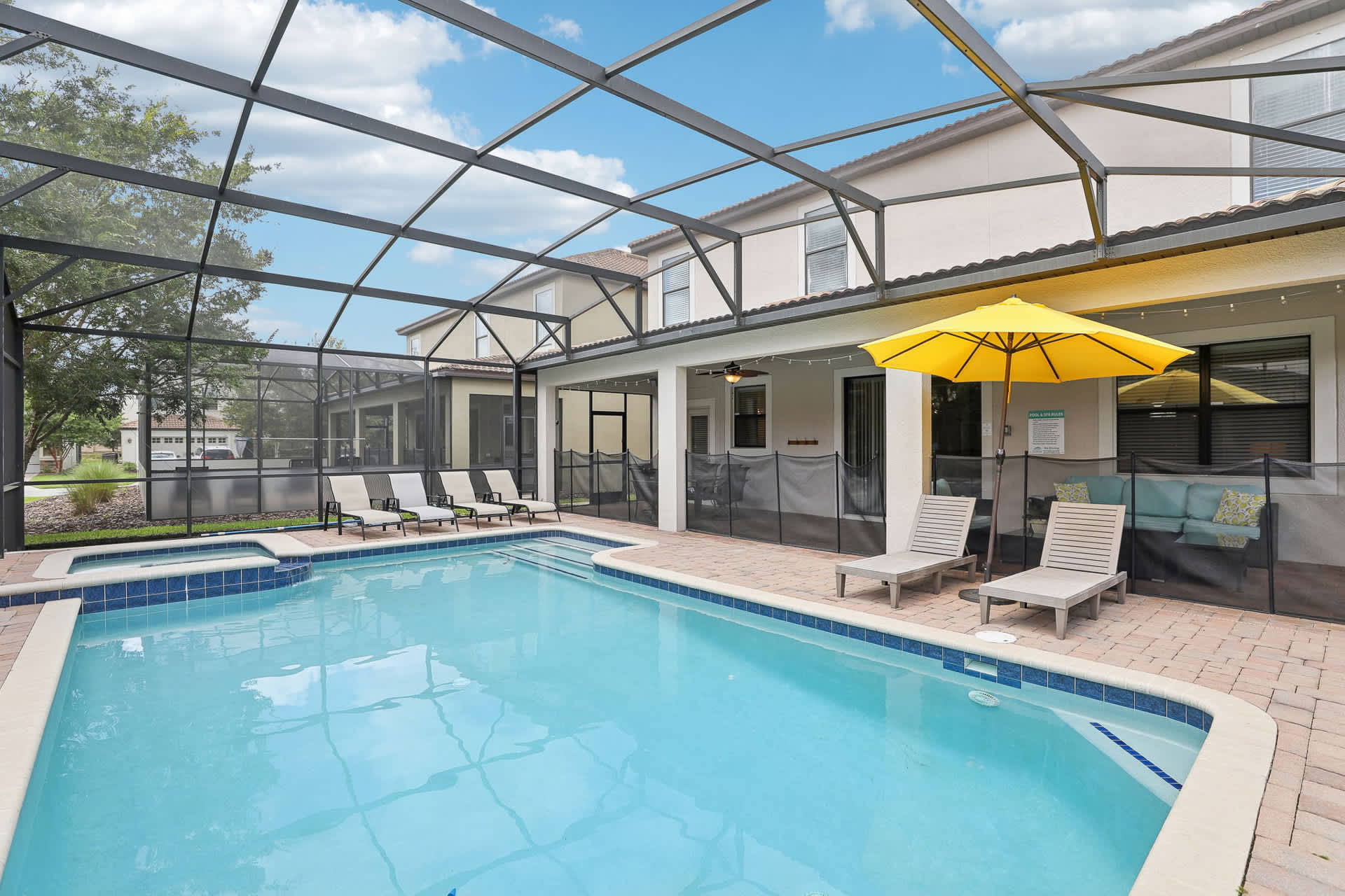 Wide view of a screened-in private pool area featuring a large swimming pool and hot tub, surrounded by brick pavers. Several lounge chairs are positioned for sunbathing, with a yellow umbrella and the back of the house visible under a bright sky.