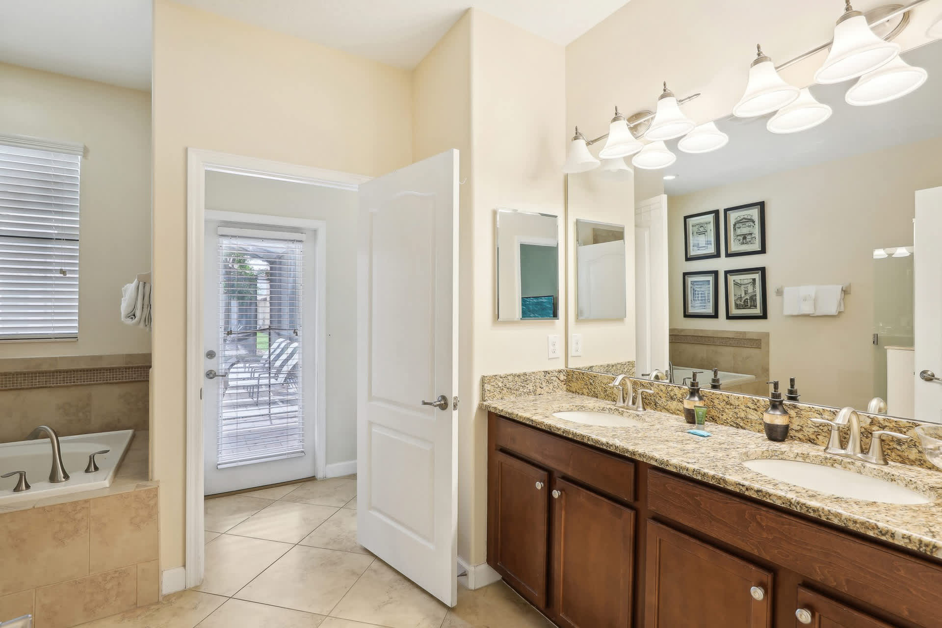 View of a spacious bathroom with a double vanity, granite countertops, and a large mirror, with an open white door revealing an outdoor patio and pool area.