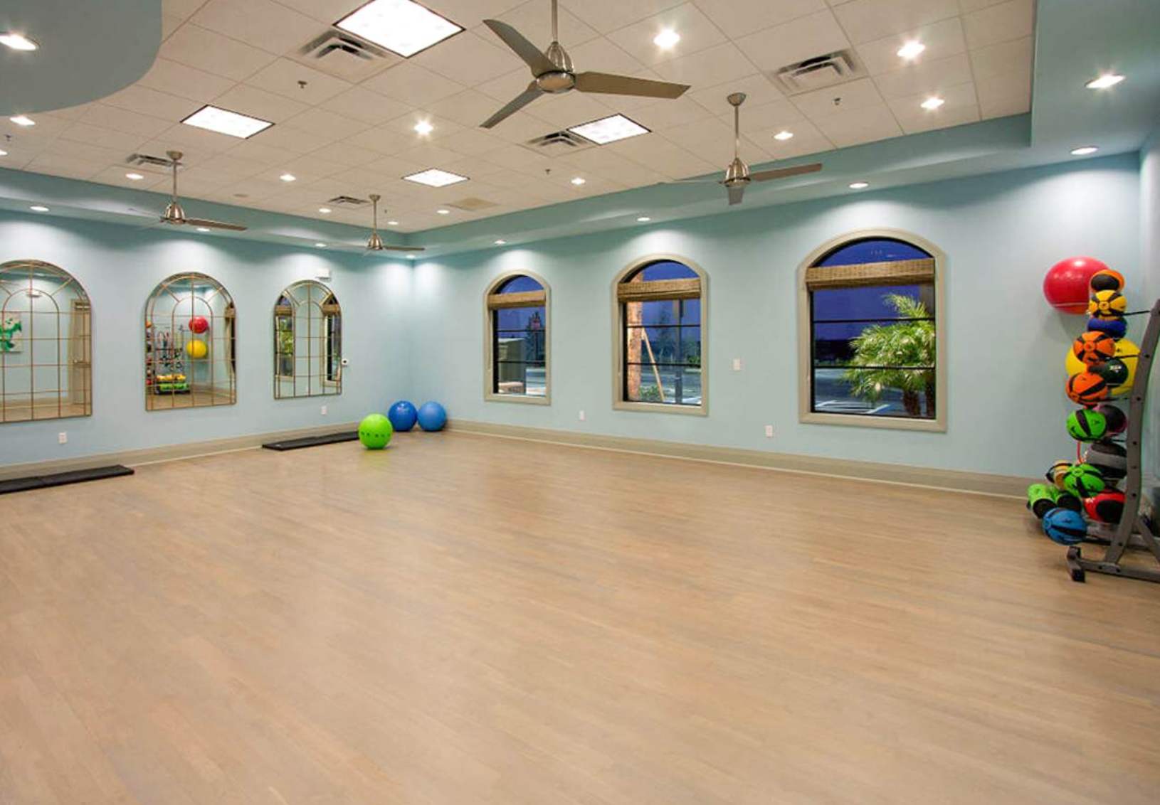 A spacious fitness room features light wood flooring, light blue walls, arched windows, multiple mirrors, and various exercise balls and weights on a rack.