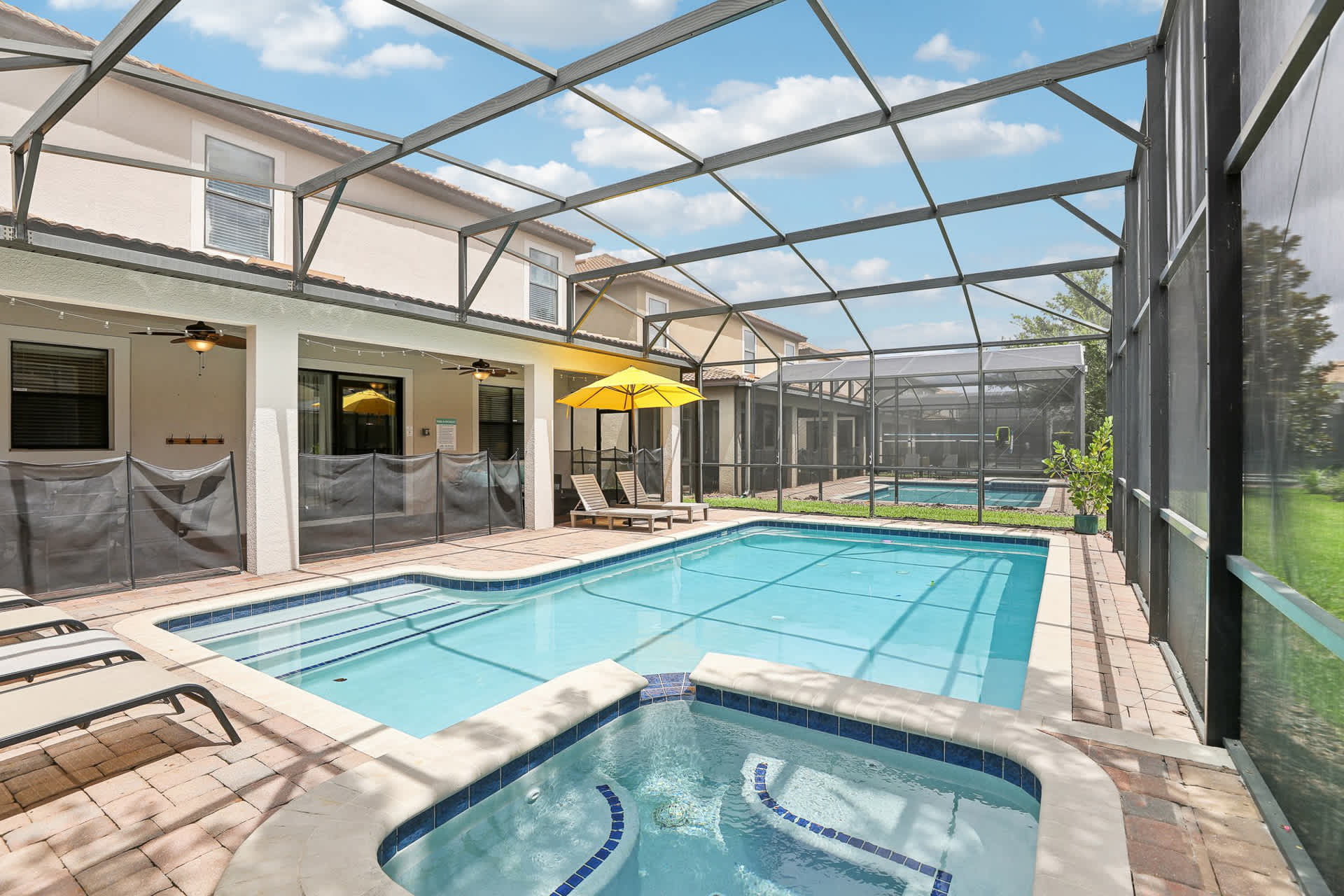 Screened-in outdoor pool area with a rectangular swimming pool and an integrated hot tub, surrounded by brick pavers. Several lounge chairs and a yellow umbrella are visible, with a safety fence separating the pool from the covered patio.