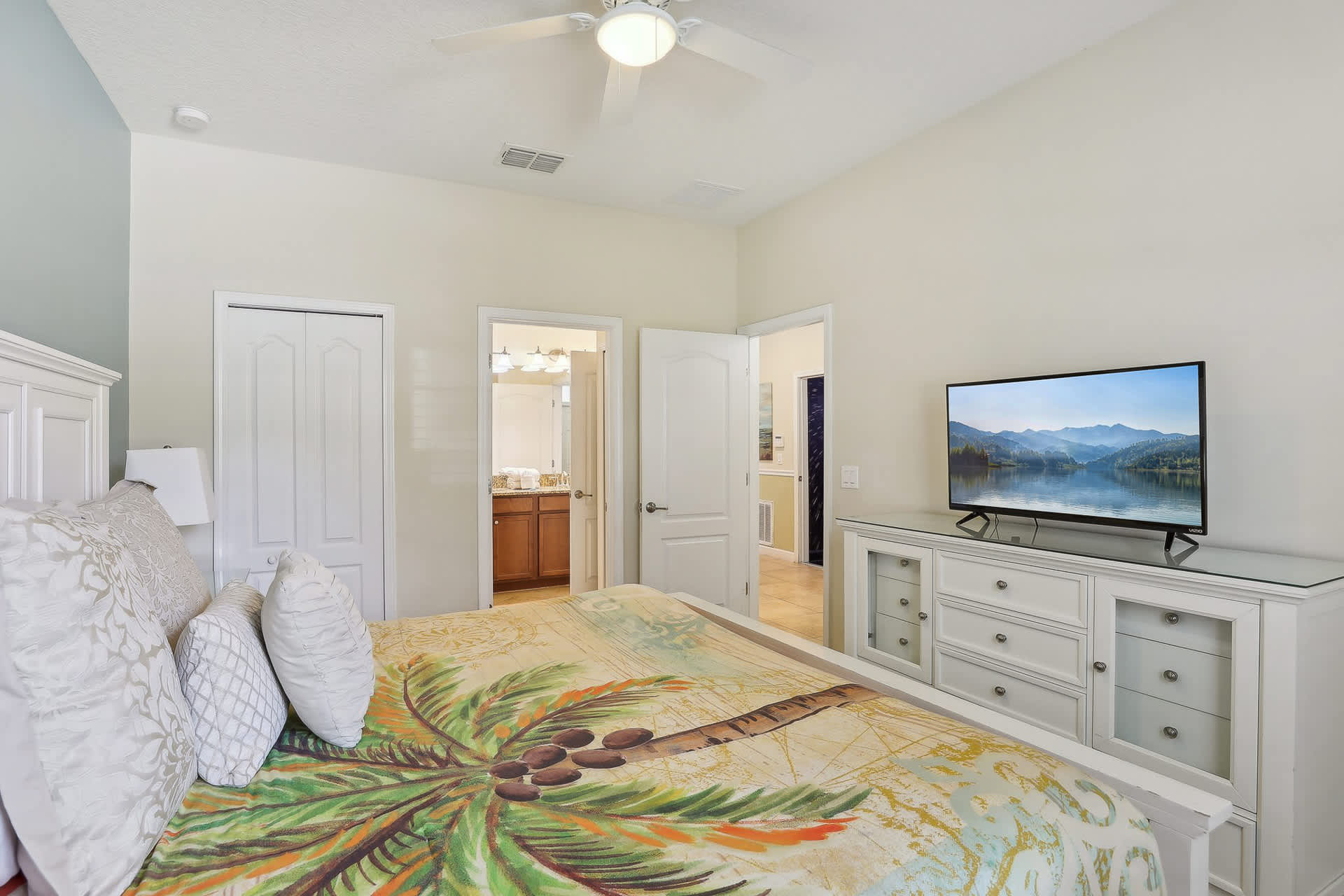 Another view of the bedroom showing a large flat-screen TV on a white dresser, a white bed with colorful bedding, and an open door leading to an en-suite bathroom.
