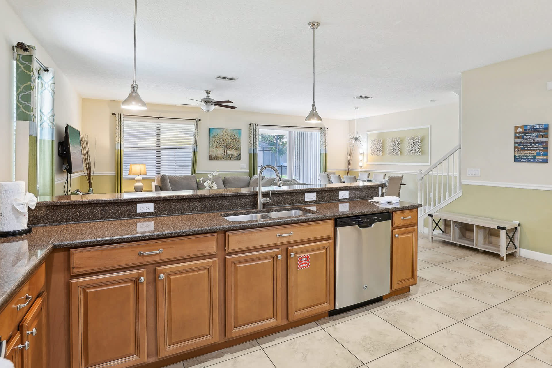 Open-concept kitchen island with a double sink and dishwasher, featuring dark granite countertops and wood cabinetry. Pendant lights hang above the breakfast bar, overlooking the living and dining areas.