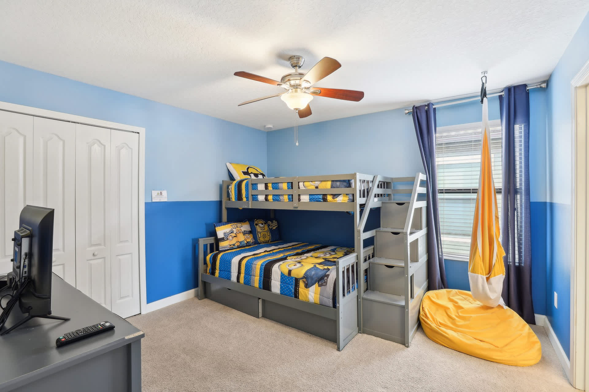 A vibrant children's bedroom features a gray bunk bed with Minions-themed bedding, blue and light blue two-tone walls, a ceiling fan, and a gray dresser with a TV.