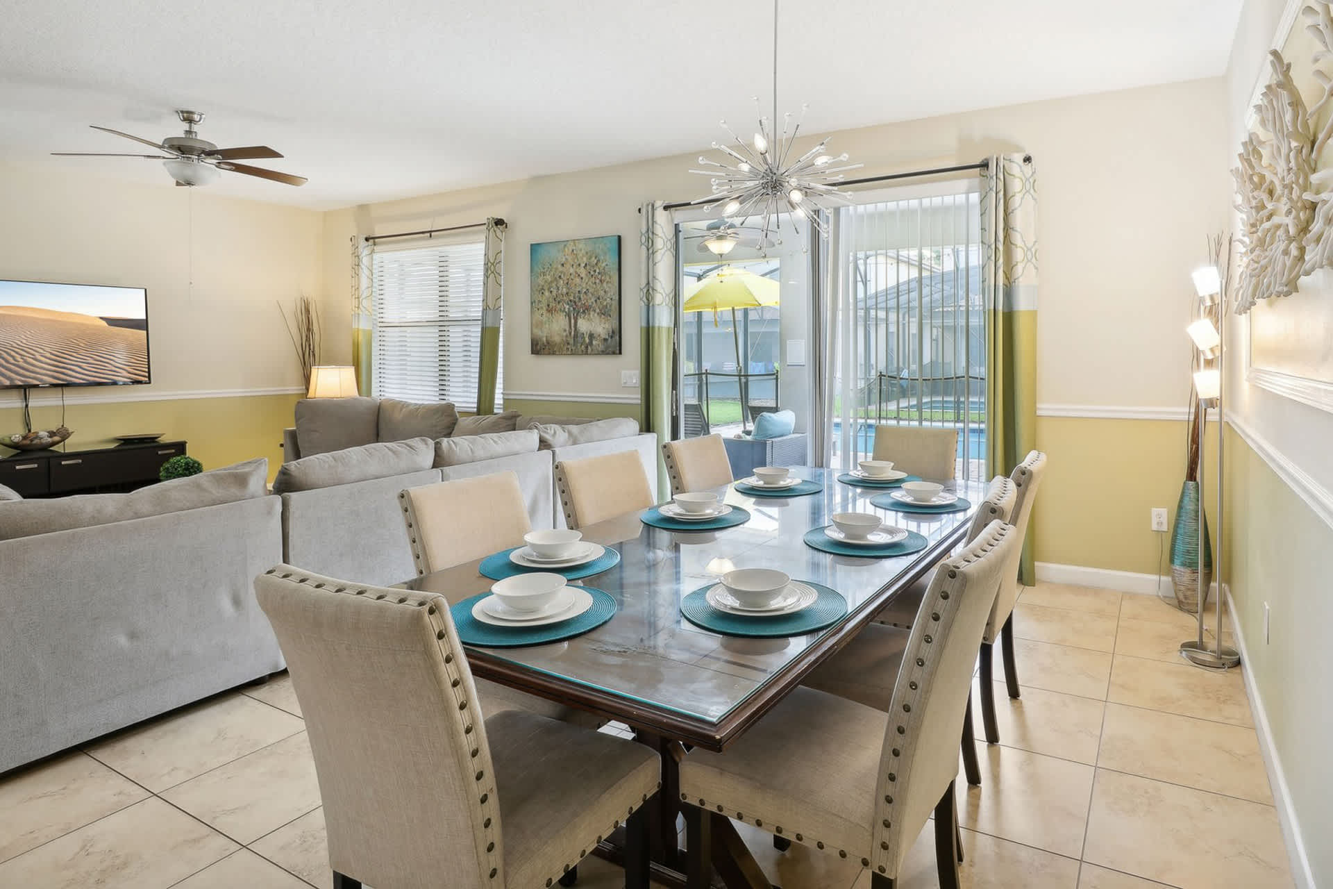 Elegant dining area with a long dark wood table set for eight, featuring light upholstered chairs and teal placemats. A modern sputnik chandelier hangs above, and sliding glass doors offer a view of the outdoor pool.