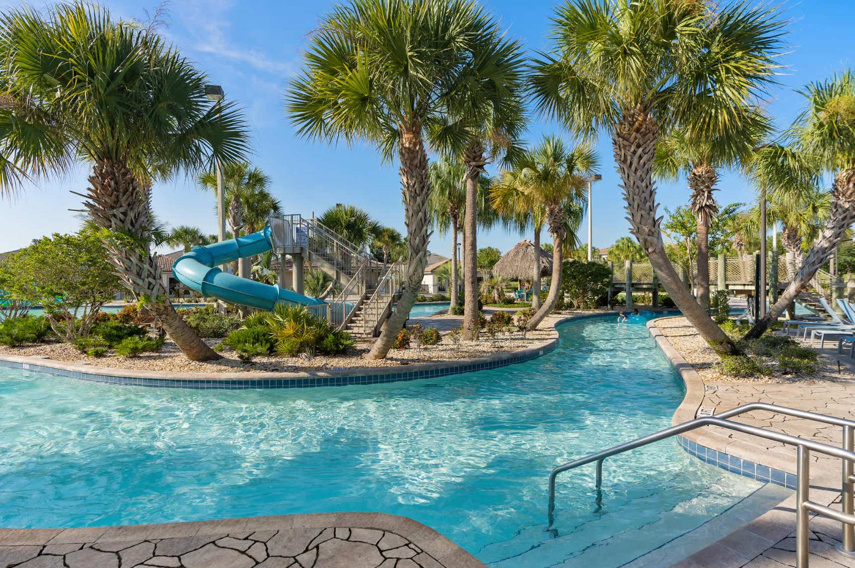 A serene lazy river pool winding through a tropical landscape, featuring a blue water slide and numerous tall palm trees. The clear blue water invites relaxation and fun.