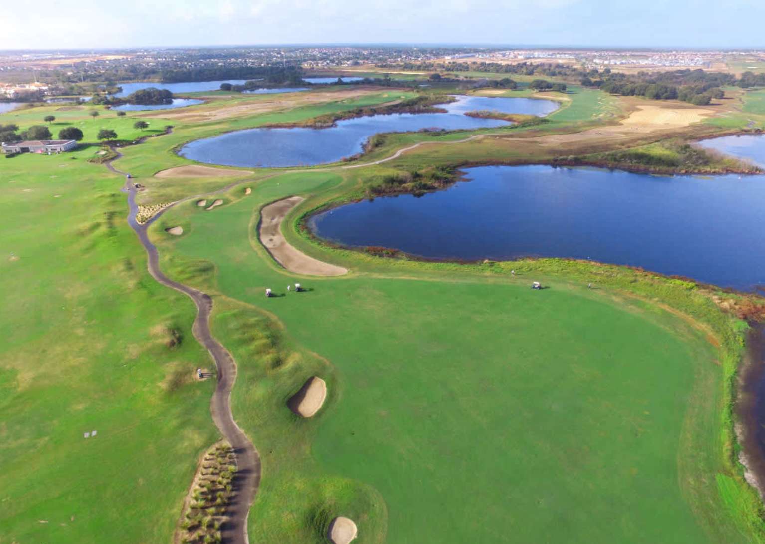 An expansive aerial view of a lush green golf course, featuring multiple large blue water hazards and winding fairways. Golf carts are visible on the course, with residential areas in the distance.