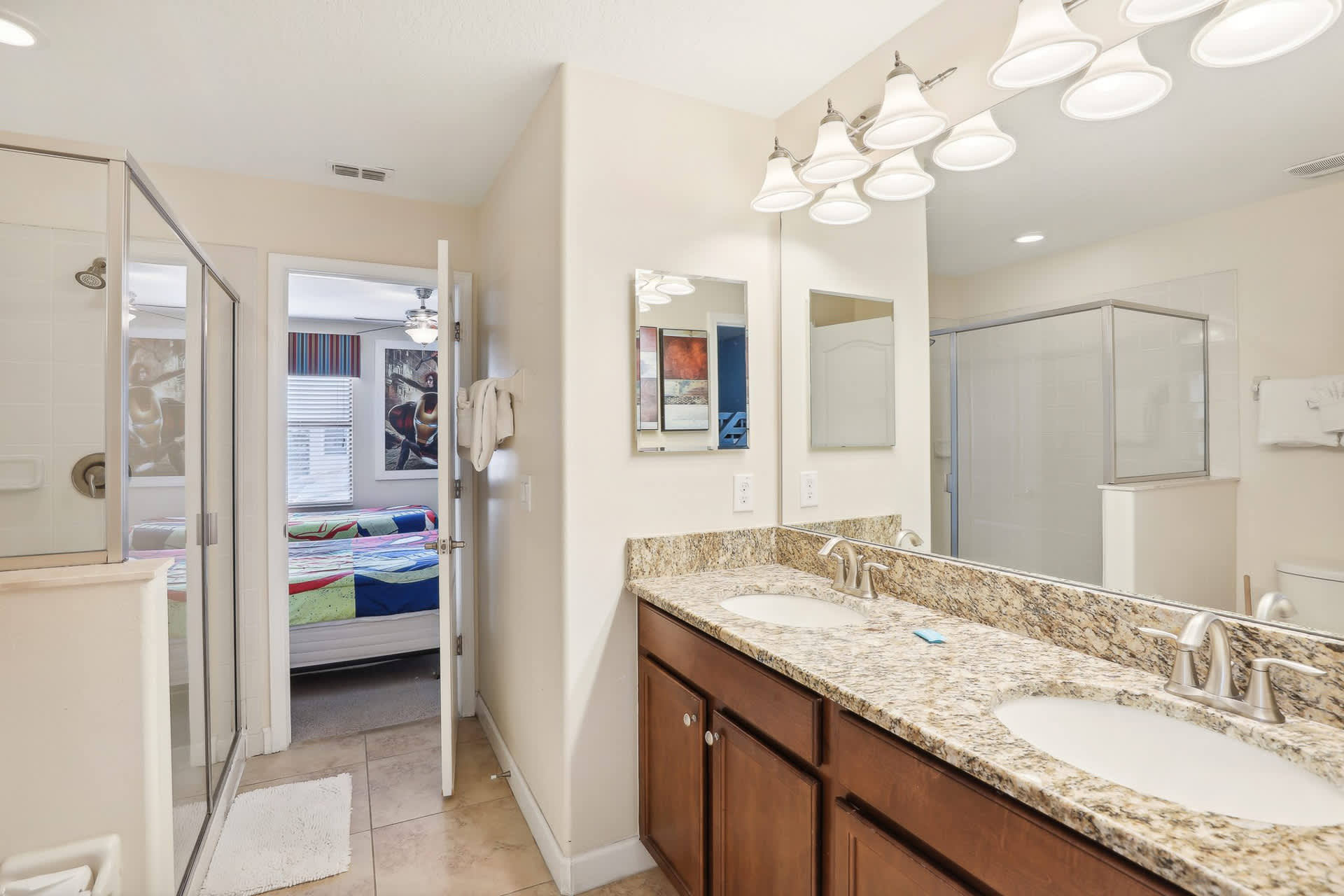 Modern bathroom with a double vanity featuring granite countertops and a large mirror, a glass-enclosed shower, and tiled flooring, with a themed bedroom visible through an open door.