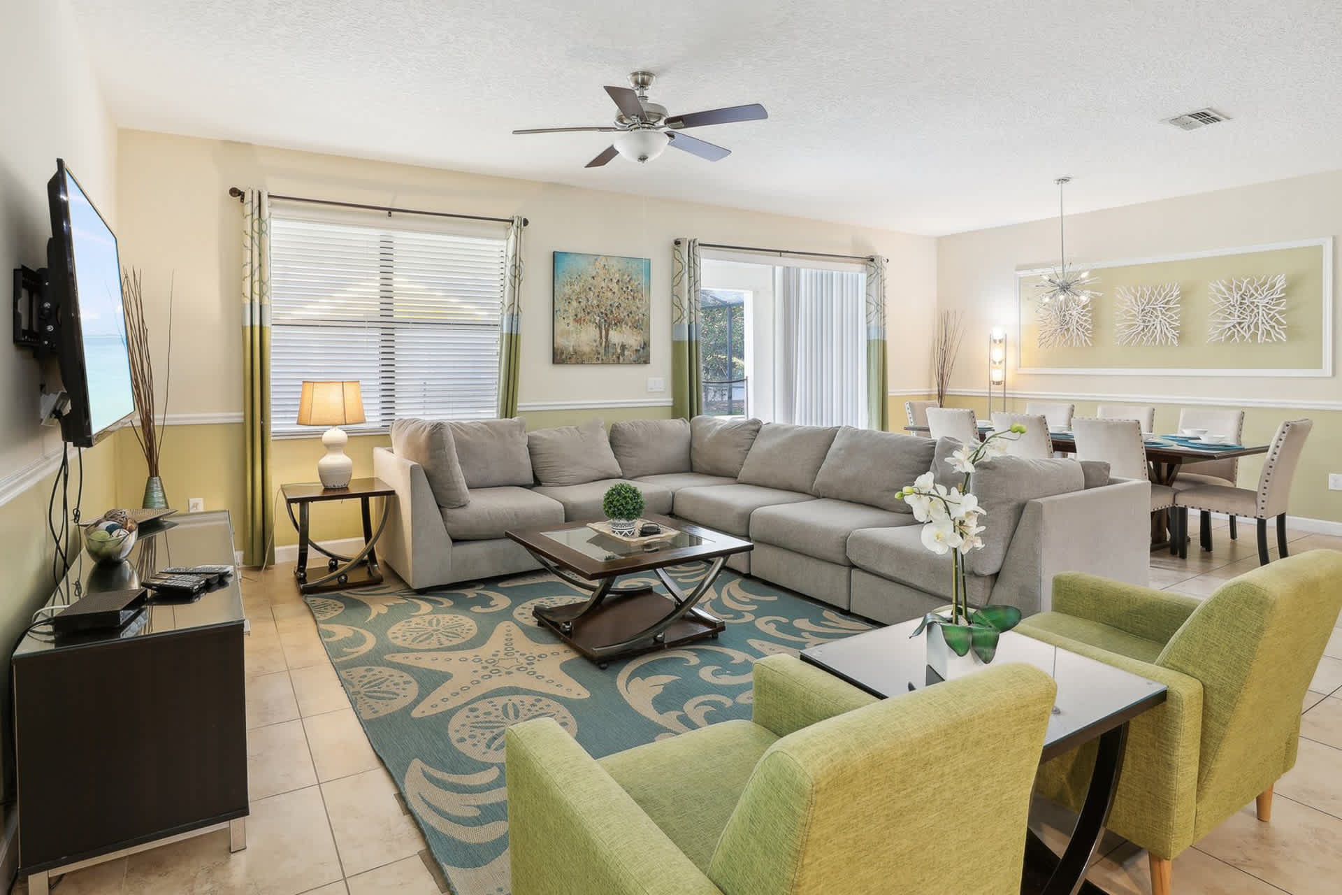 Bright living room featuring a large gray sectional sofa, a dark wood coffee table, and two lime green armchairs on a blue and white patterned rug. A flat-screen TV is mounted on the wall, and a dining area with a long table is visible in the background.
