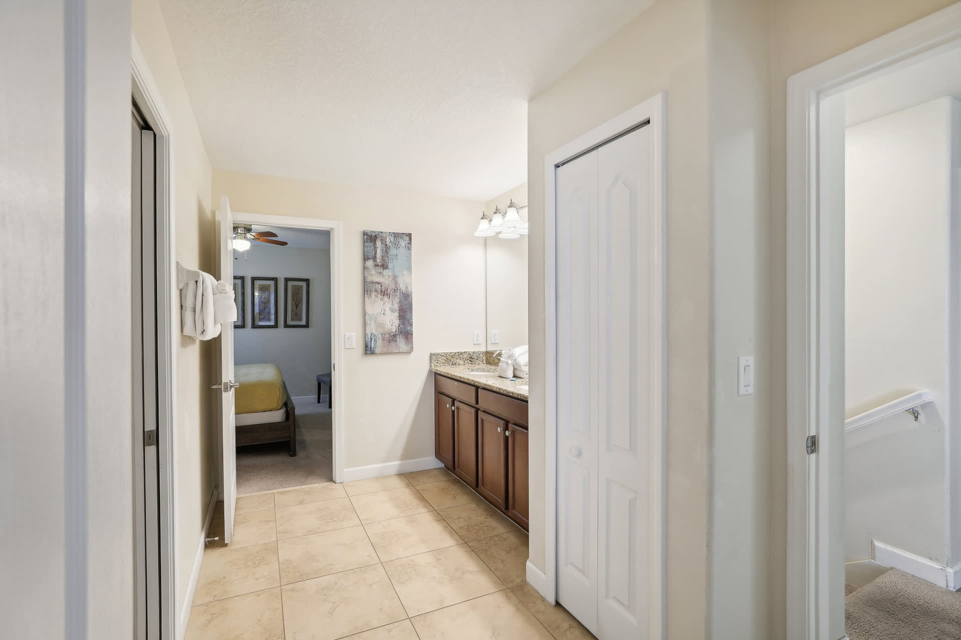 Hallway with light tiled flooring leading into a bathroom with a partial view of a double vanity, and an open doorway revealing another bedroom.