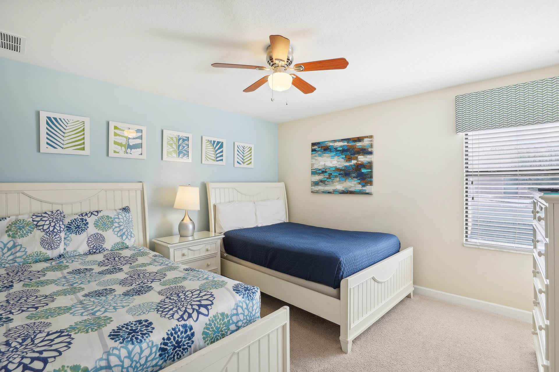 Kids' bedroom with two white beds, one with blue floral bedding and the other with a solid blue comforter, a light blue accent wall, and a ceiling fan with wooden blades.