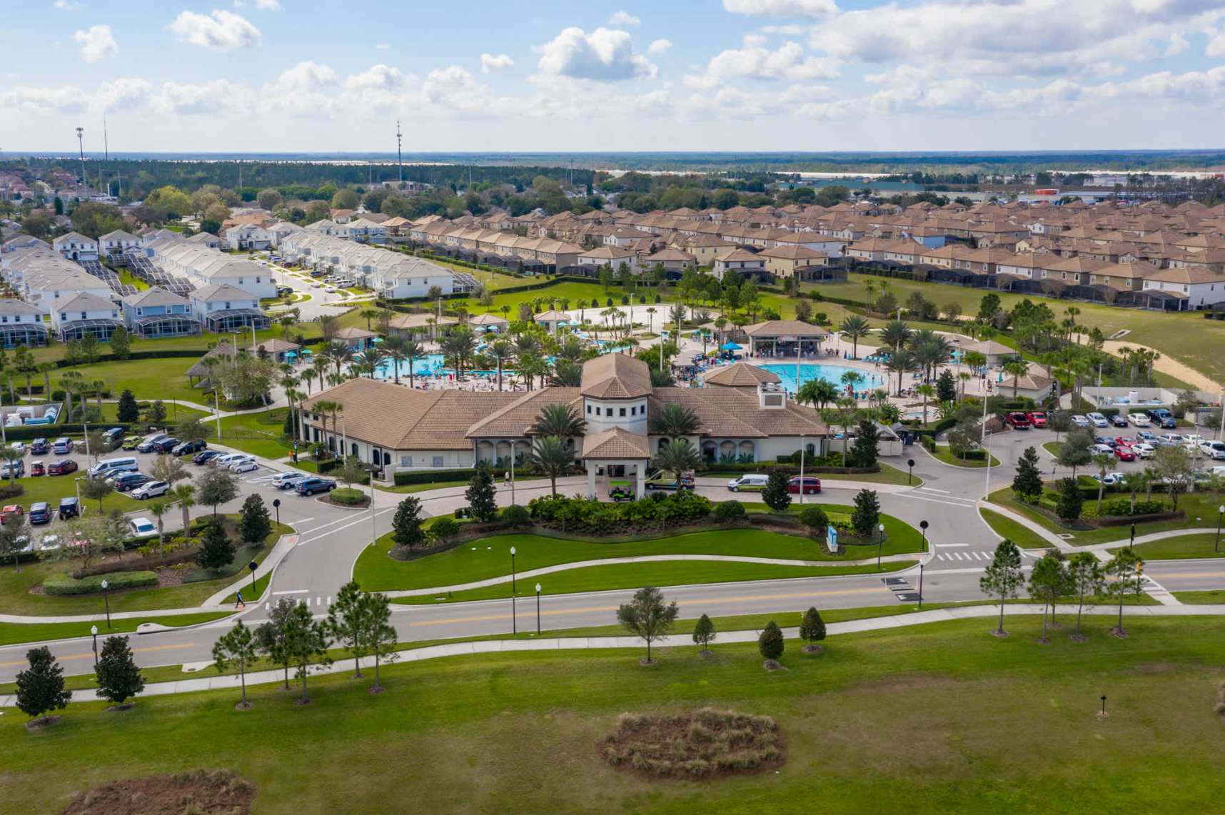 An aerial view shows a large clubhouse building with a circular driveway, surrounded by lush landscaping, and a resort-style communal pool complex with numerous lounge chairs and palm trees.