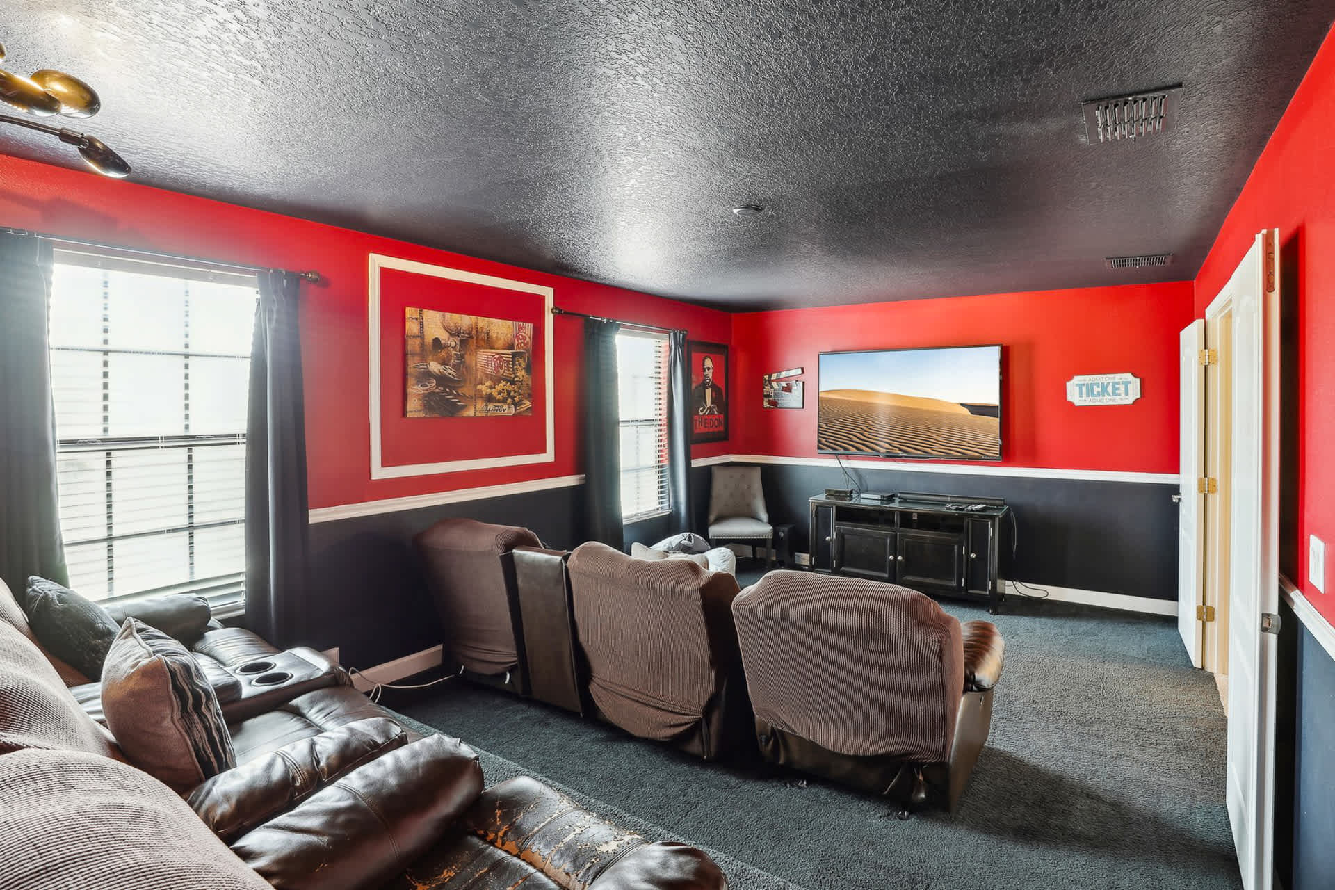 Cozy media room with red and black two-tone walls, featuring multiple rows of brown reclining cinema seats facing a large wall-mounted flat-screen TV. Dark carpet covers the floor, and windows are covered with dark drapes.