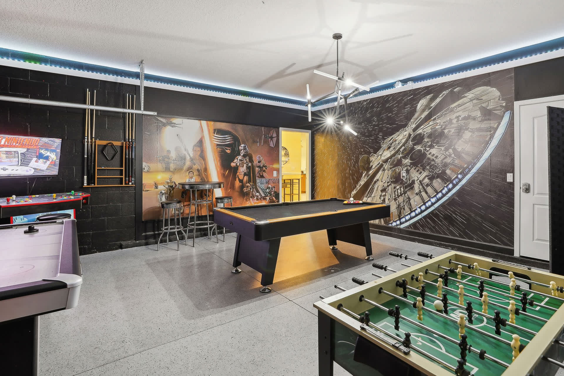 Star Wars-themed game room with a black pool table as the centerpiece, flanked by a foosball table and arcade machines. The walls feature large murals of Star Wars characters and spacecraft, with black and gray painted sections.