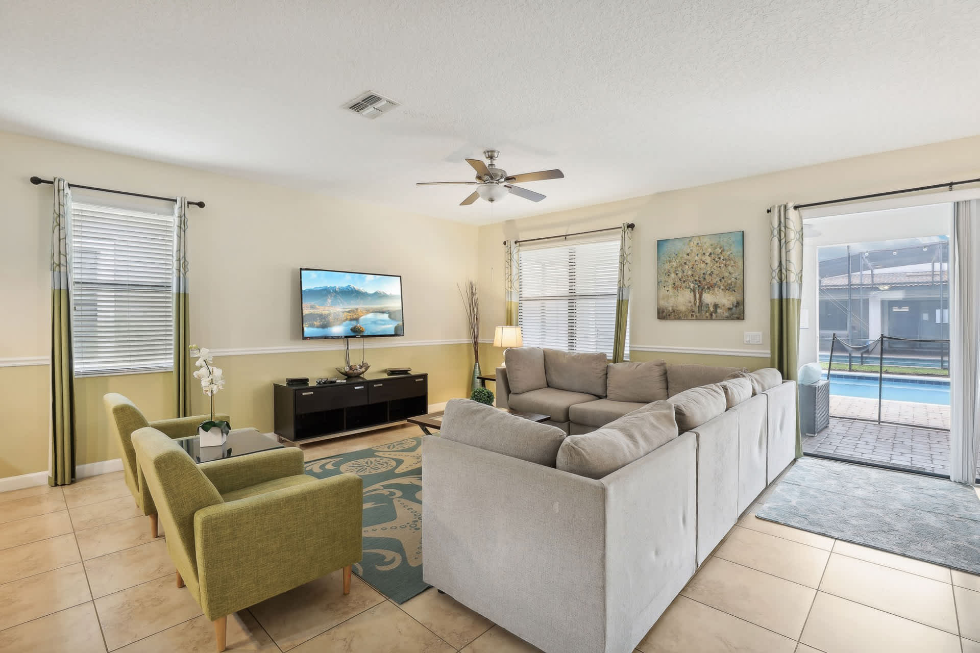 Spacious living room with a large light gray sectional sofa facing a wall-mounted TV above a dark media console. Sliding glass doors lead to an outdoor pool area, and two green armchairs are visible to the left.