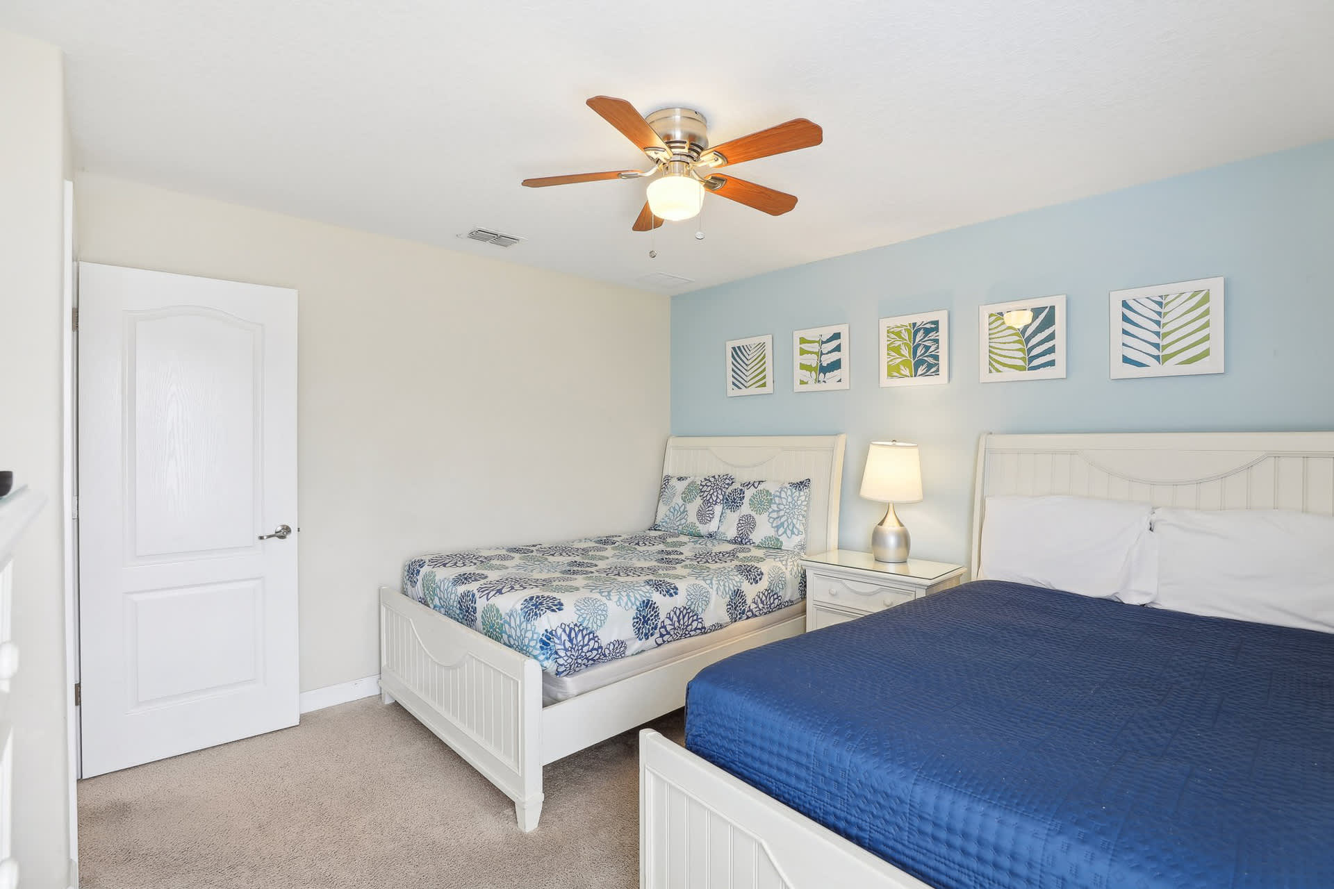 Kids' bedroom with two white beds, one with blue floral bedding and the other with a solid blue comforter, a light blue accent wall with framed botanical prints, and a ceiling fan.