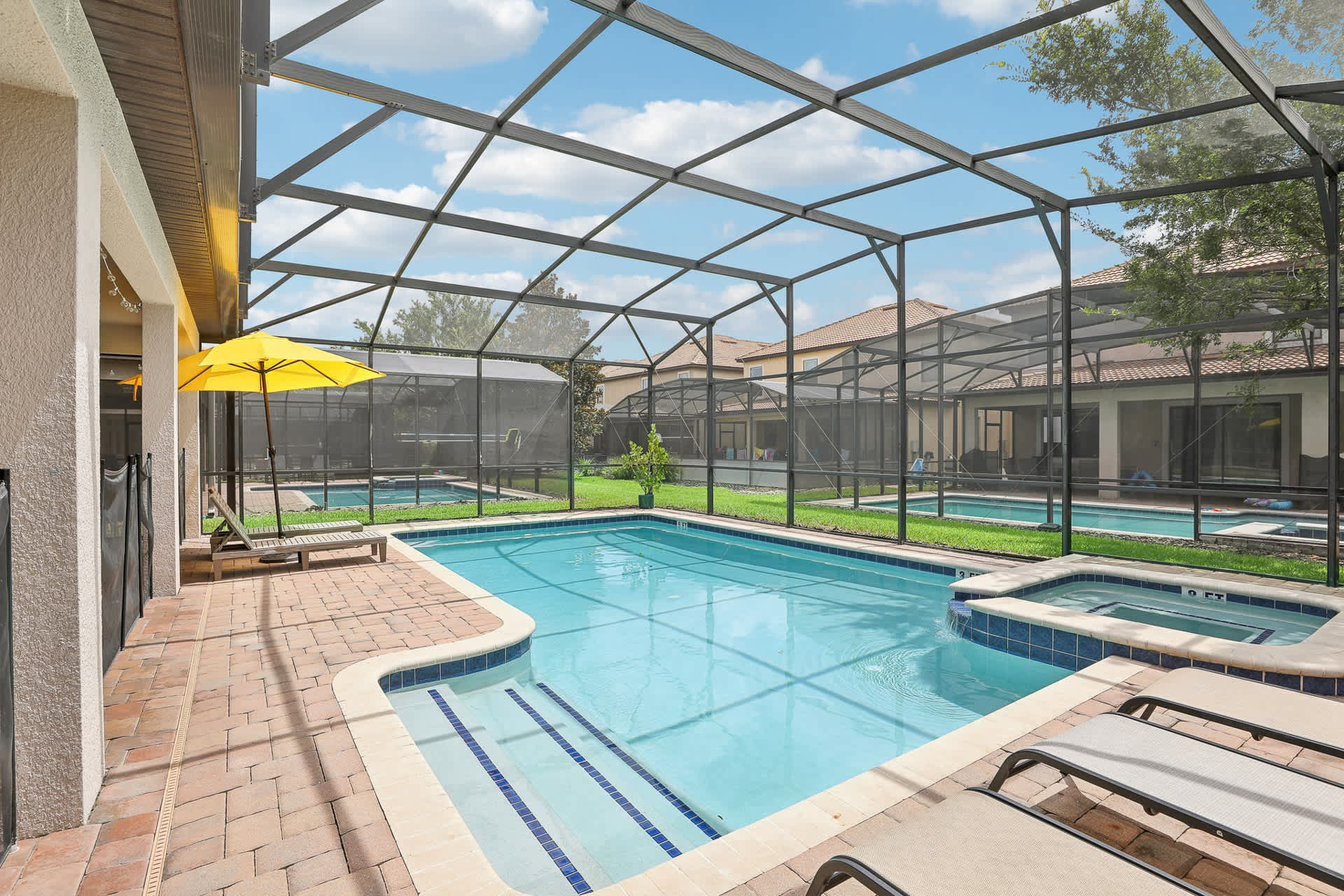 Expansive screened-in pool area with a large swimming pool and integrated hot tub, surrounded by brick pavers. Multiple lounge chairs are arranged around the pool, with a yellow umbrella providing shade, and the house's back facade visible.