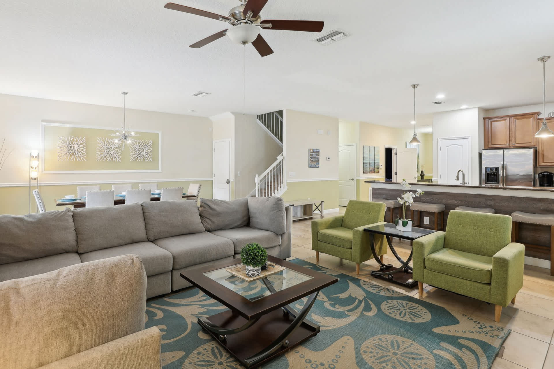 Bright open-concept living room featuring a large gray sectional sofa, two green armchairs, and a dark wood coffee table on a teal and beige patterned rug. A ceiling fan hangs above, and a dining area is visible in the background.