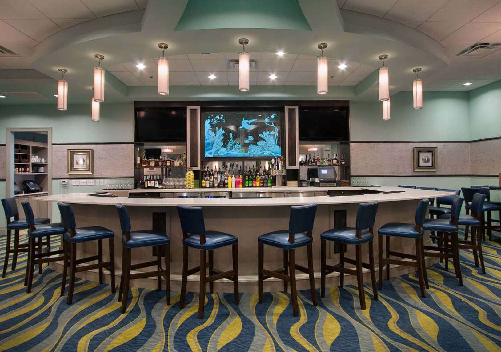 A stylish bar area with an oval counter, blue leather bar stools, and a back wall featuring shelves of liquor bottles and a large screen displaying an aquatic scene. The floor is covered with a distinctive blue and yellow wavy carpet.
