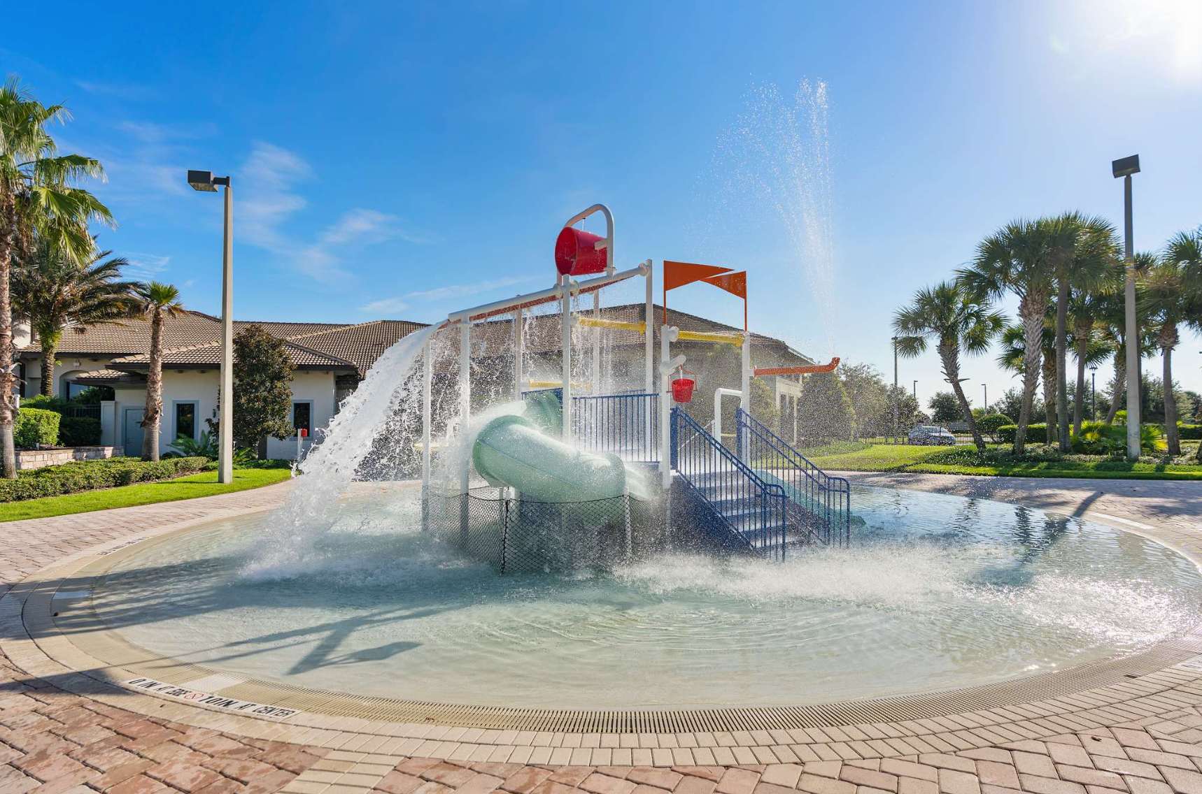 A vibrant children's splash pad featuring a large tipping bucket, a small water slide, and various water jets. The circular play area is surrounded by a brick patio and palm trees under a clear blue sky.