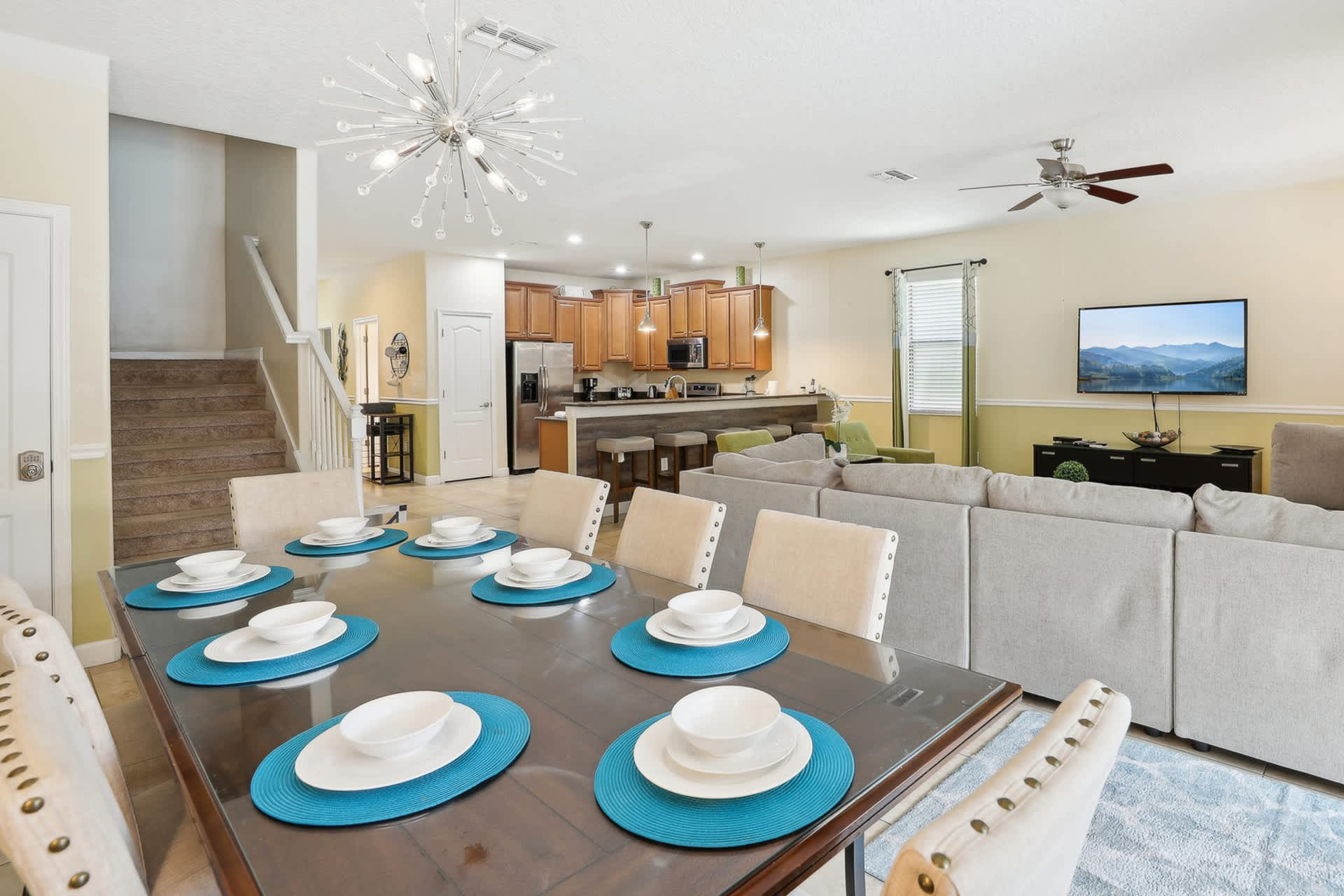 View of the dining area with a large table set for eight, looking towards the open-concept kitchen and living room. A modern chandelier illuminates the space, and a staircase is visible in the background.