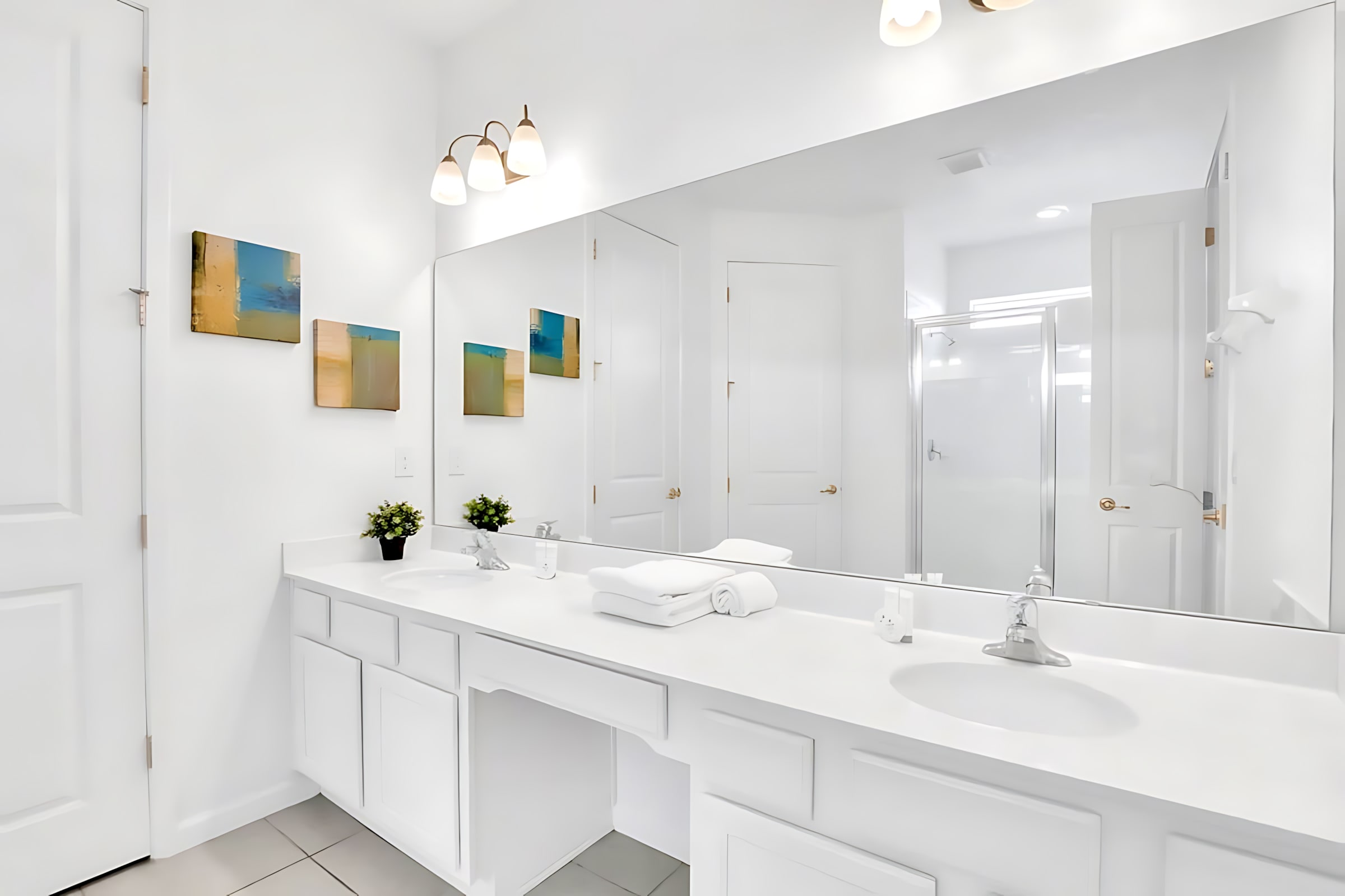 Spacious white bathroom featuring a double vanity with white cabinets and countertop, a large mirror spanning the wall, and abstract wall art. A shower enclosure is visible in the mirror's reflection.