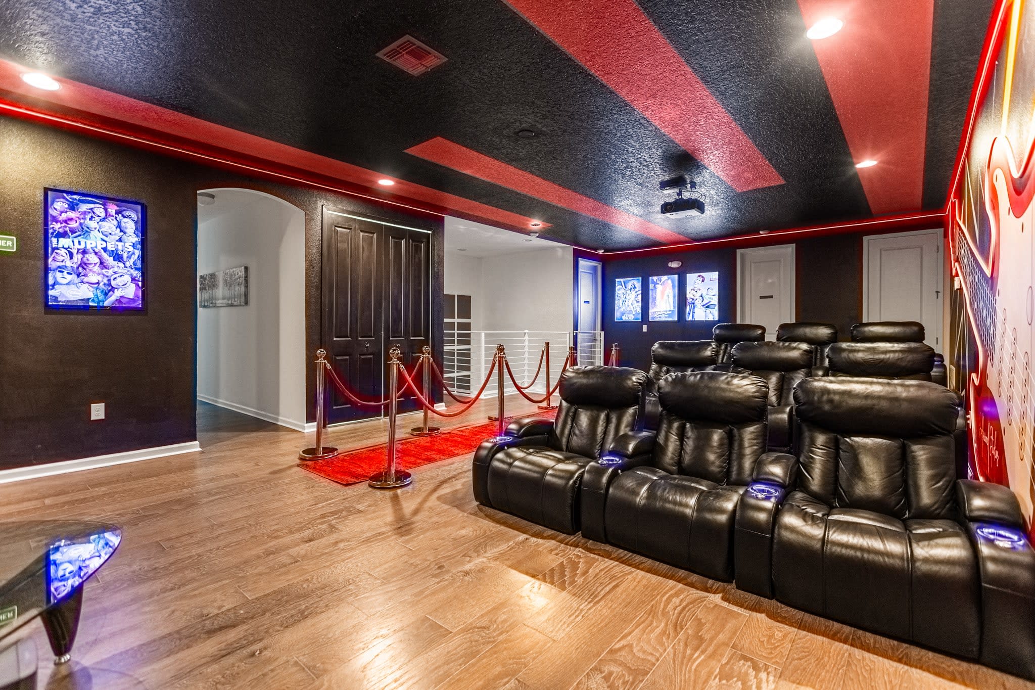 Wide shot of the home theater room, showing multiple rows of black leather reclining seats facing a large projection screen. Red velvet ropes guide the entrance, and movie posters adorn the walls.