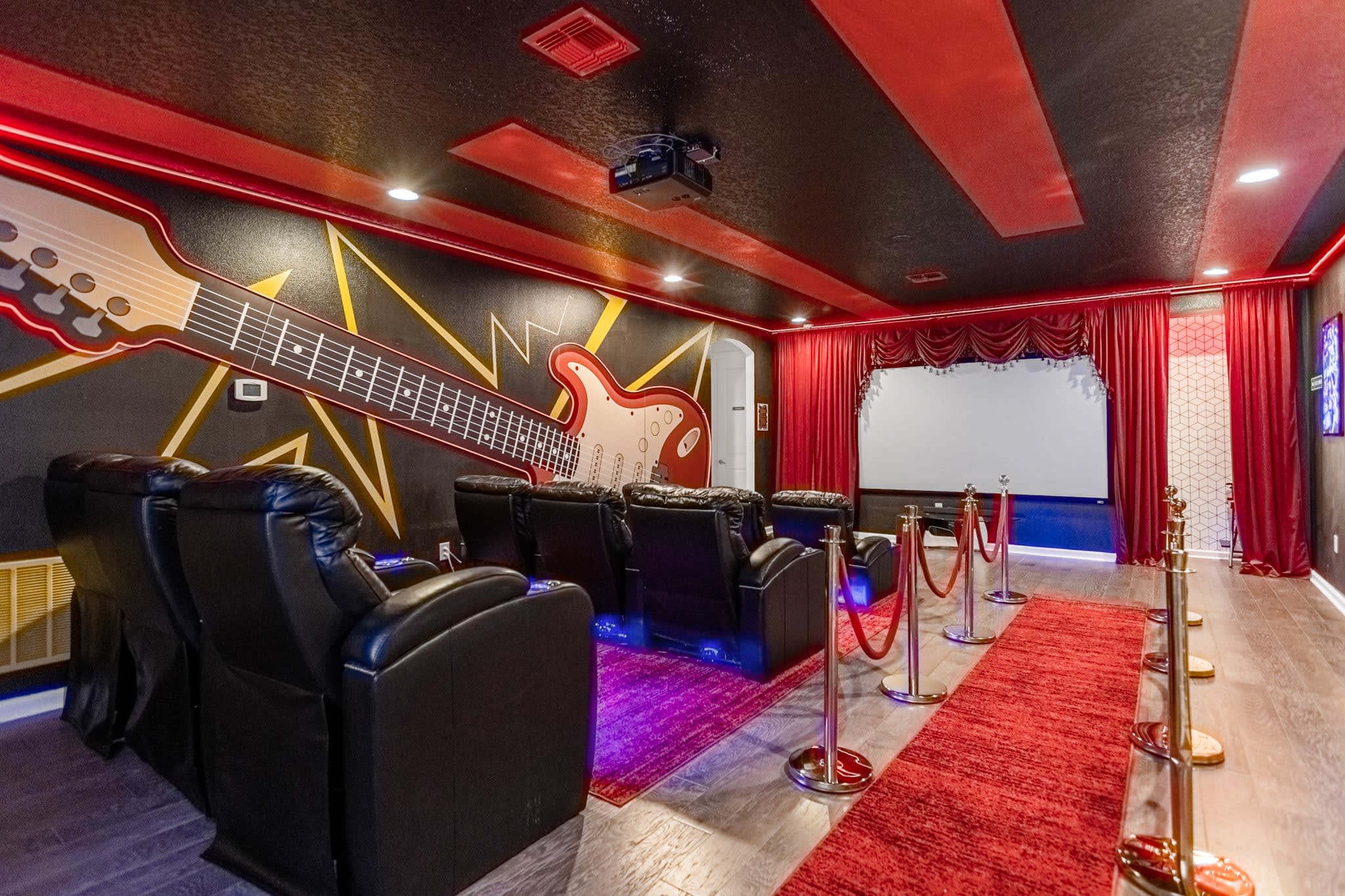 Themed home theater room with black reclining seats, a large projection screen, and red velvet curtains. The walls feature a large guitar mural and red accent lighting, with red carpet leading to the screen.