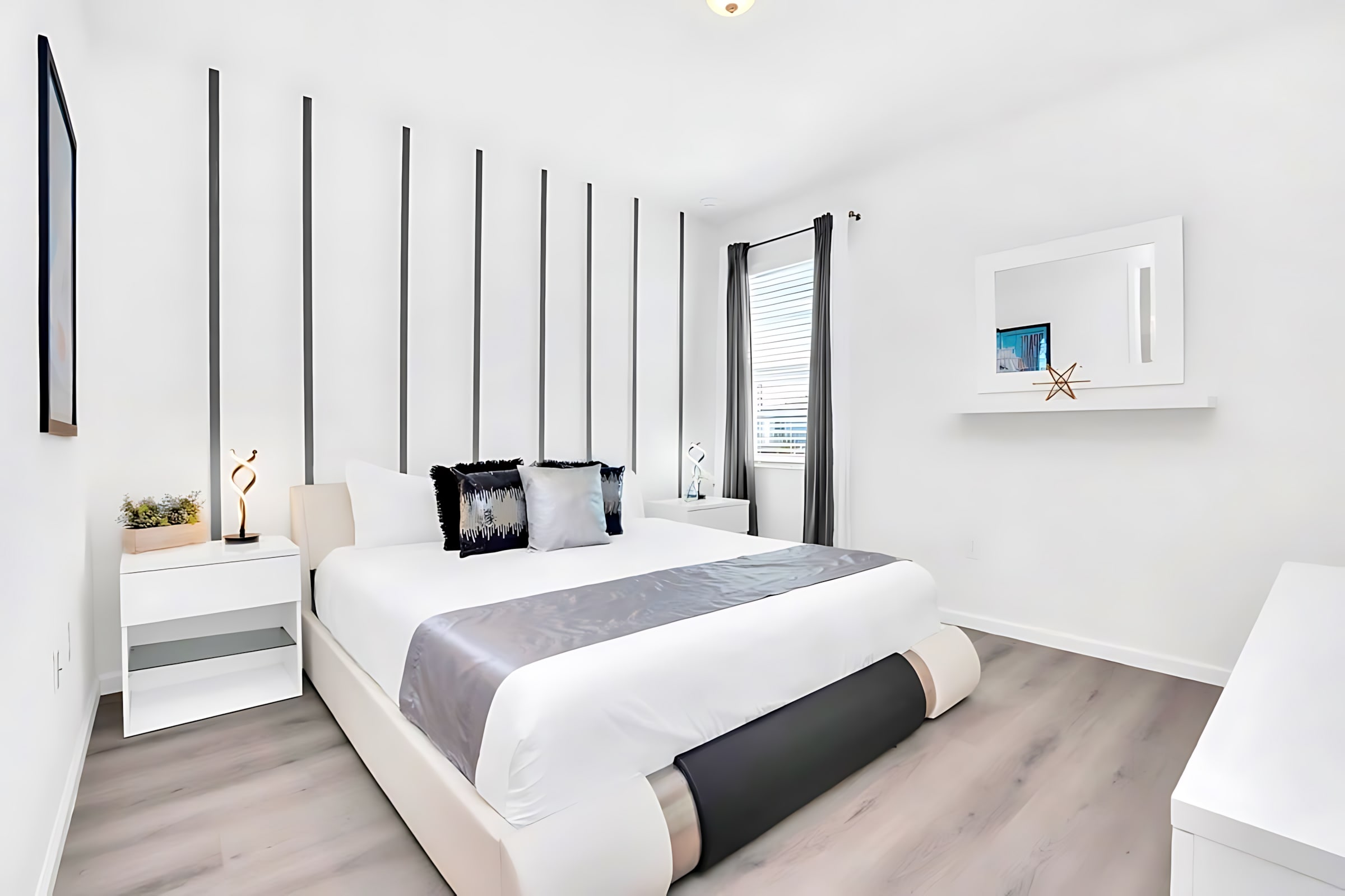 A contemporary bedroom with a king-sized bed, featuring a striking white and black vertical striped accent wall behind the headboard. White nightstands with modern lamps flank the bed, and a window with dark curtains provides natural light.