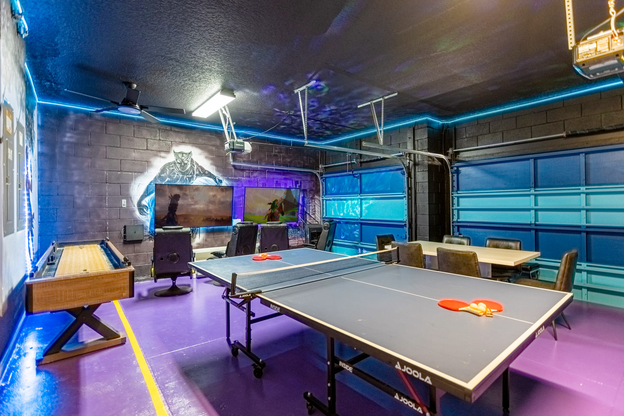 Another view of the vibrant game room, highlighting a shuffleboard table, a ping pong table, and multiple large flat-screen TVs. The room features a purple floor, blue LED lighting, and garage doors with blue accents.