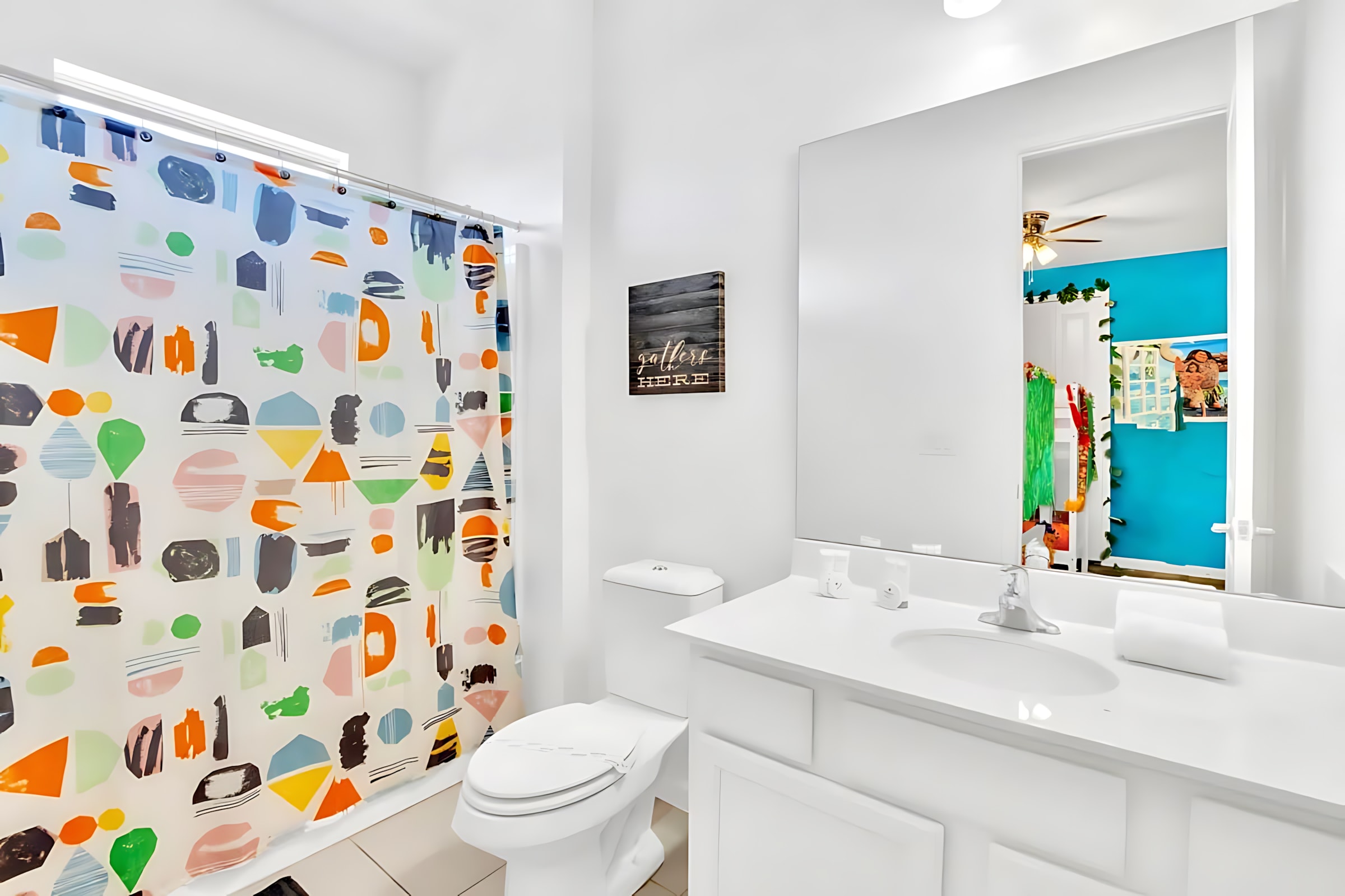 Bright white bathroom with a single vanity, toilet, and a shower featuring a colorful geometric-patterned curtain. The mirror reflects the blue Moana-themed bedroom.