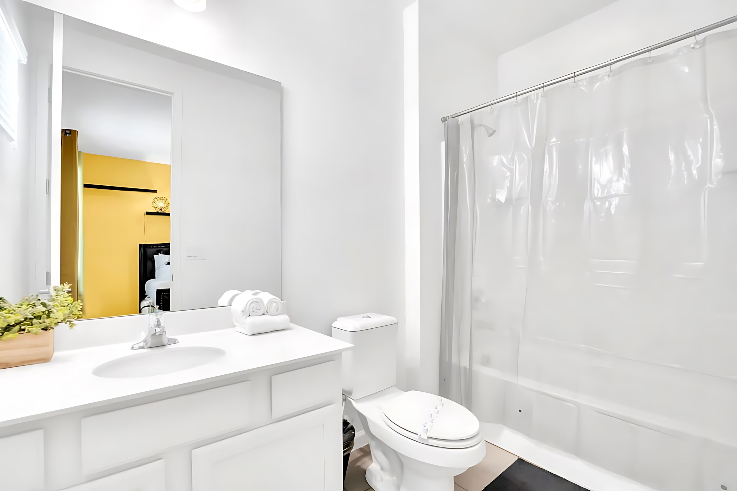 Simple white bathroom with a single vanity, large mirror, toilet, and a shower/tub combo with a clear shower curtain, offering a view into the adjacent yellow bedroom.
