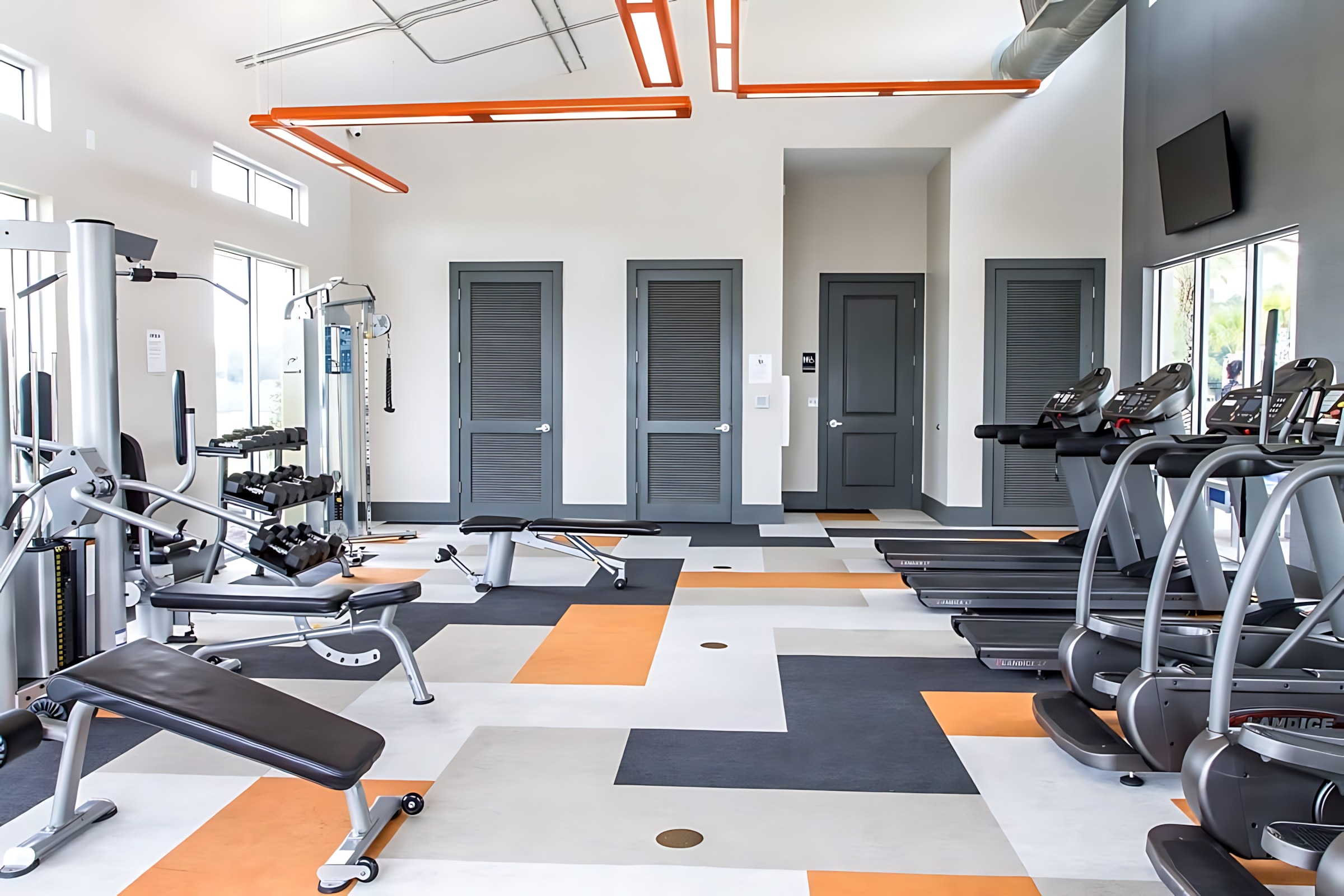 Modern fitness center equipped with various exercise machines including treadmills, ellipticals, and weight benches, set on a vibrant geometric floor with orange and gray patterns. The space features high ceilings with exposed ductwork, linear orange lighting, and large windows providing ample natural light.