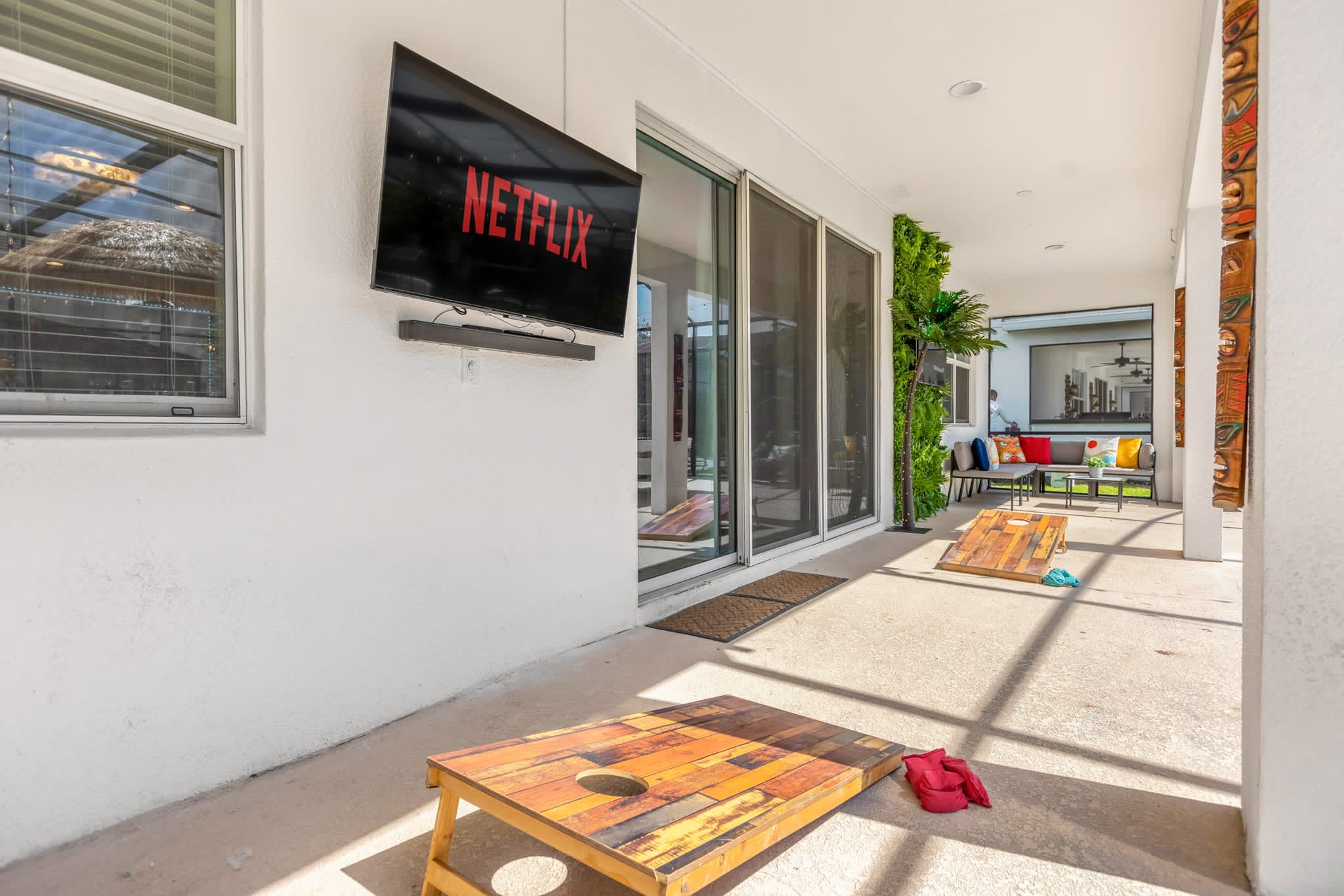 Covered outdoor patio area with two cornhole boards and a wall-mounted TV displaying the Netflix logo. Sliding glass doors lead into the house, and tropical-themed decor is visible in the background.
