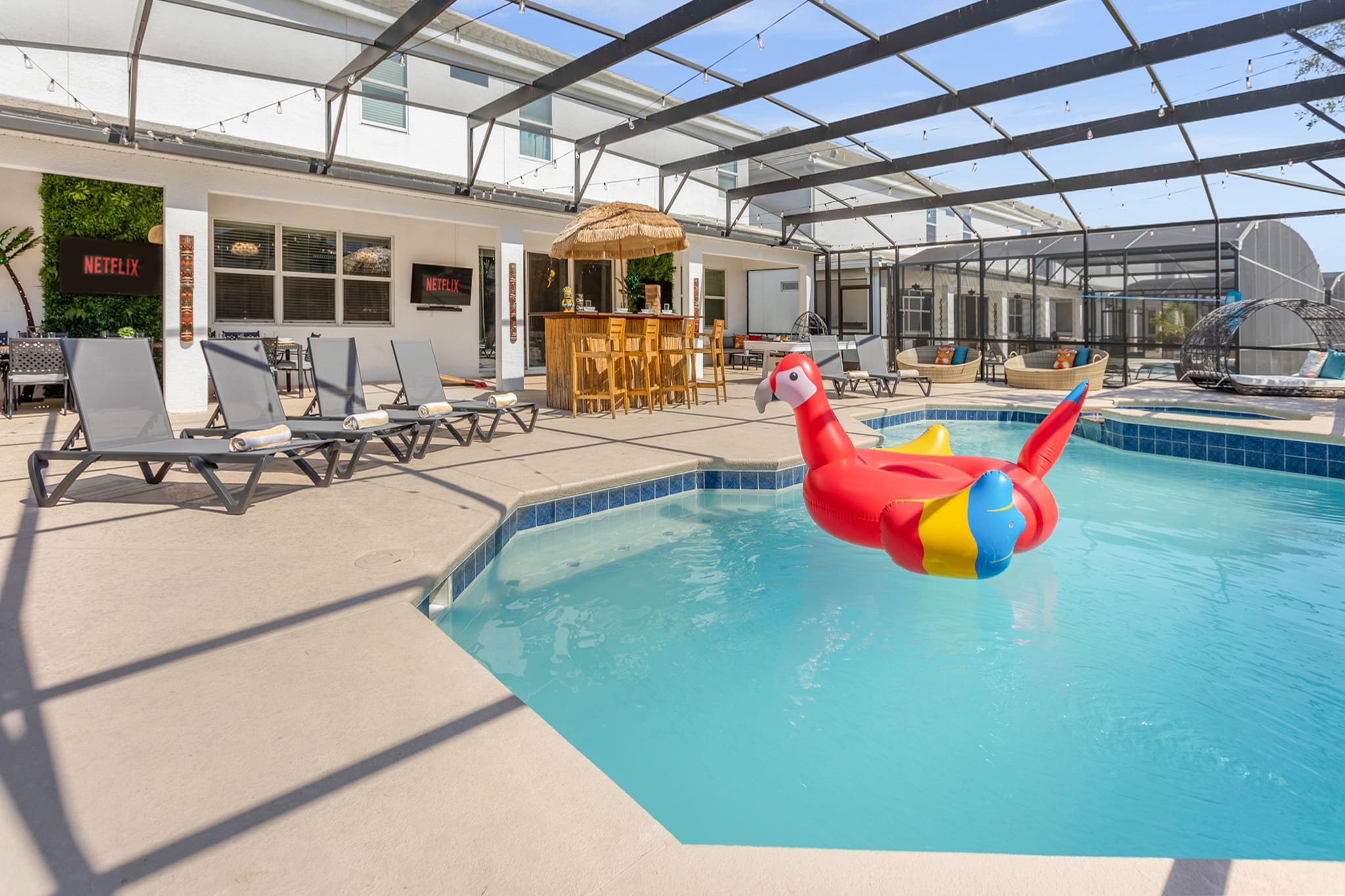 Screened-in outdoor pool area featuring a large swimming pool with a red parrot float, several lounge chairs, and a tiki bar with stools. String lights are strung across the enclosure, and a wall-mounted TV is visible near the bar.