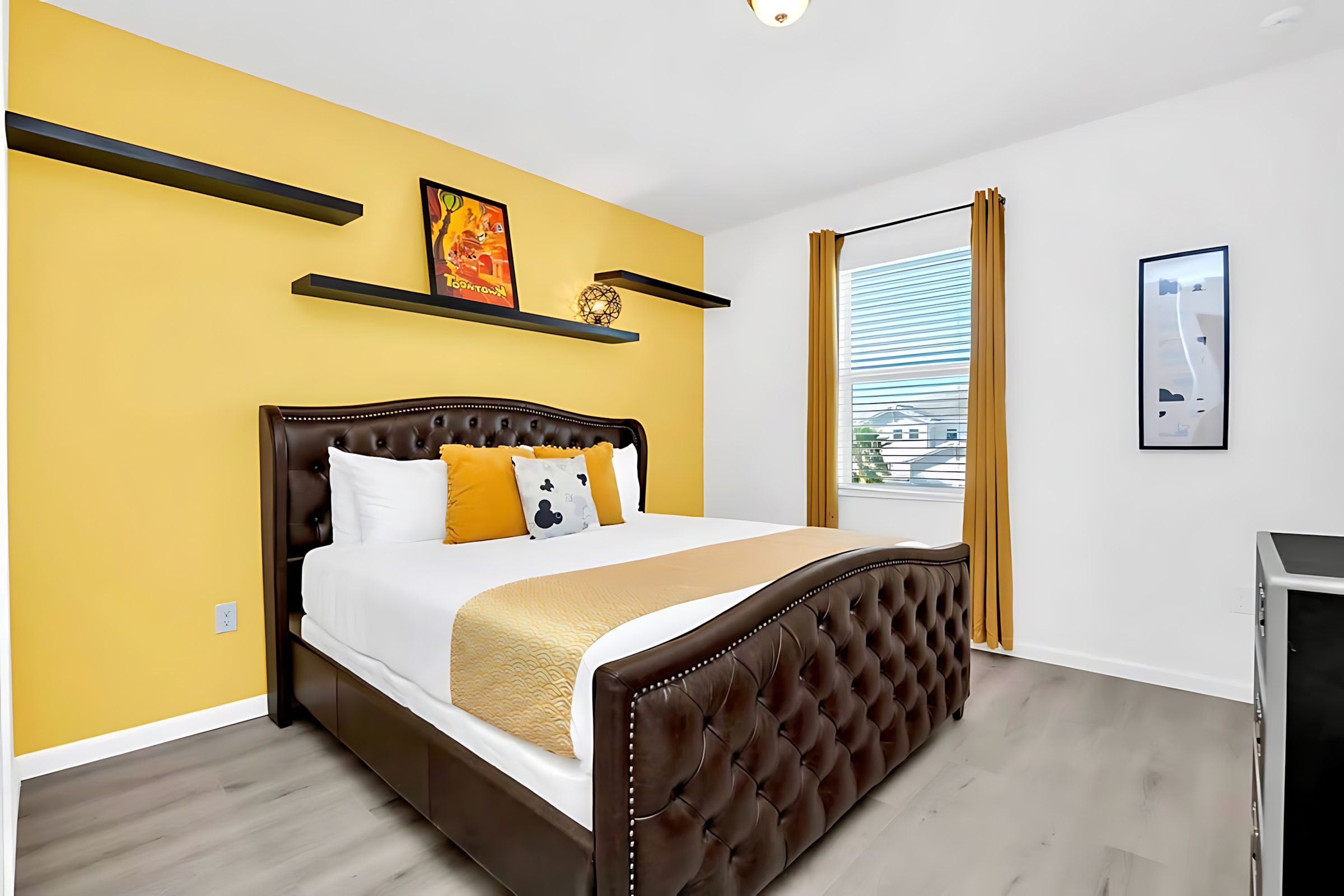 Warm bedroom featuring a tufted brown leather king-sized bed, white bedding, and a sunny yellow accent wall with black floating shelves and a window.