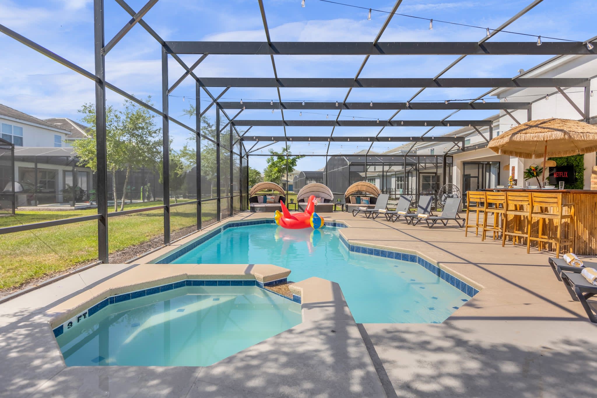 Wide view of a screened-in outdoor pool area, showcasing a rectangular swimming pool with an attached hot tub and a red parrot float. Several lounge chairs and covered daybeds are arranged around the pool, with string lights overhead.
