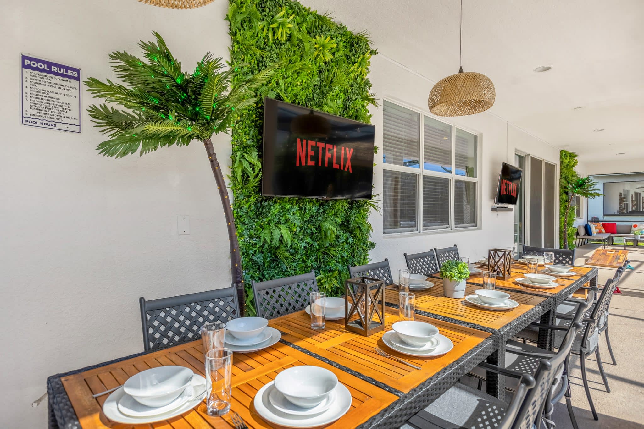 Covered outdoor dining area on a patio, featuring a long wooden table set for a meal with black chairs. A large wall-mounted TV is set against a vibrant green artificial plant wall, next to a decorative palm tree.